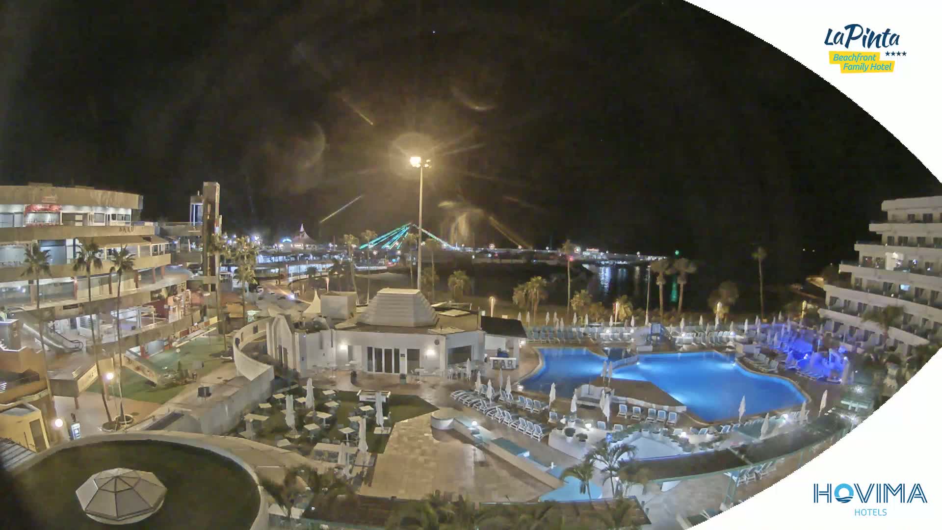 An illuminated resort pool area at night overlooks a calm body of water and a bridge under a partly cloudy sky.