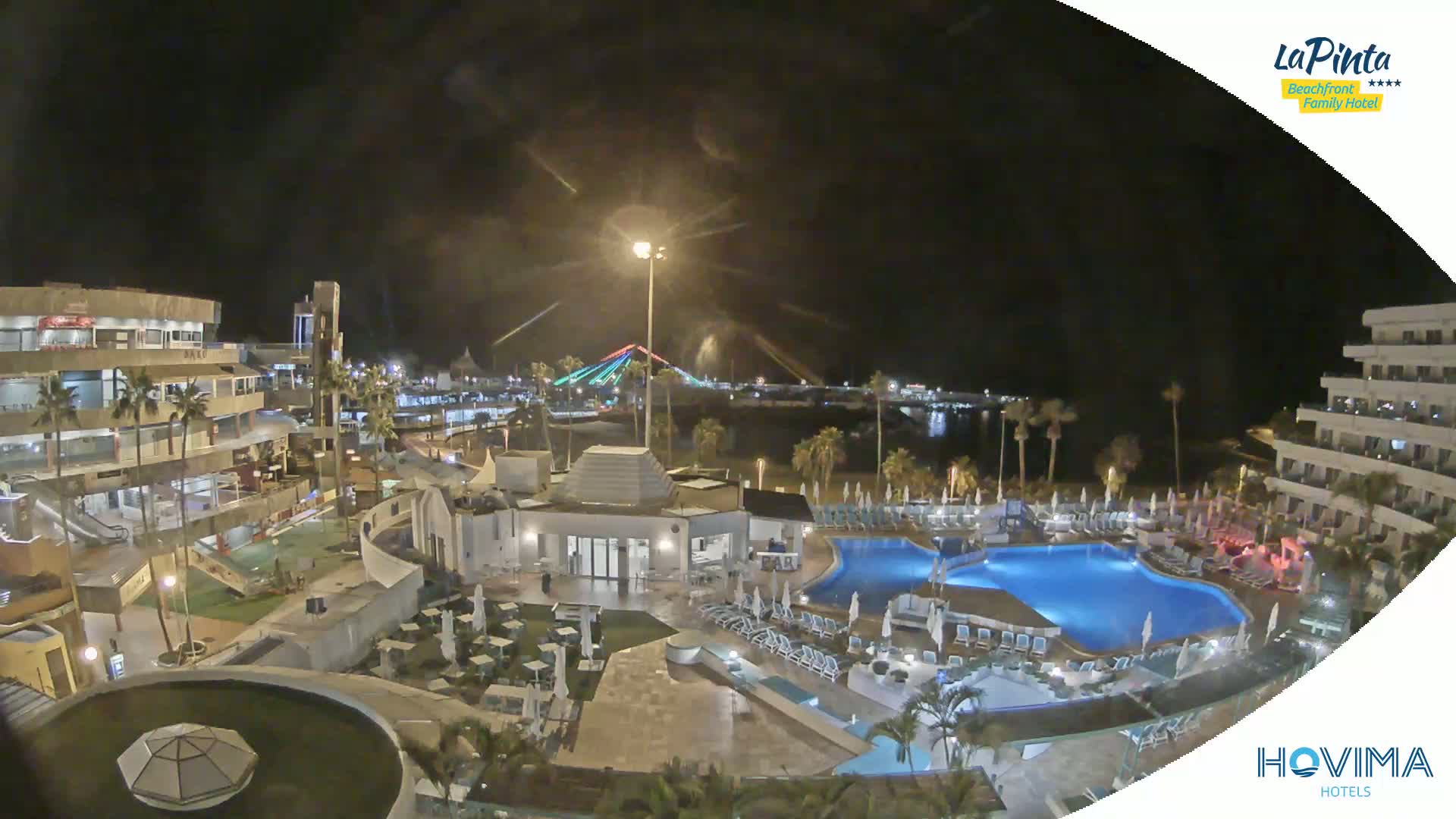 A nighttime aerial view of a resort complex with swimming pools and surrounding buildings, illuminated under a dark sky.