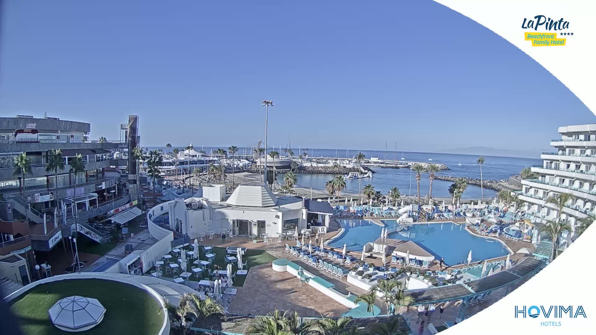 A resort pool area with surrounding buildings and a marina is seen on a sunny day, overlooking a calm ocean.