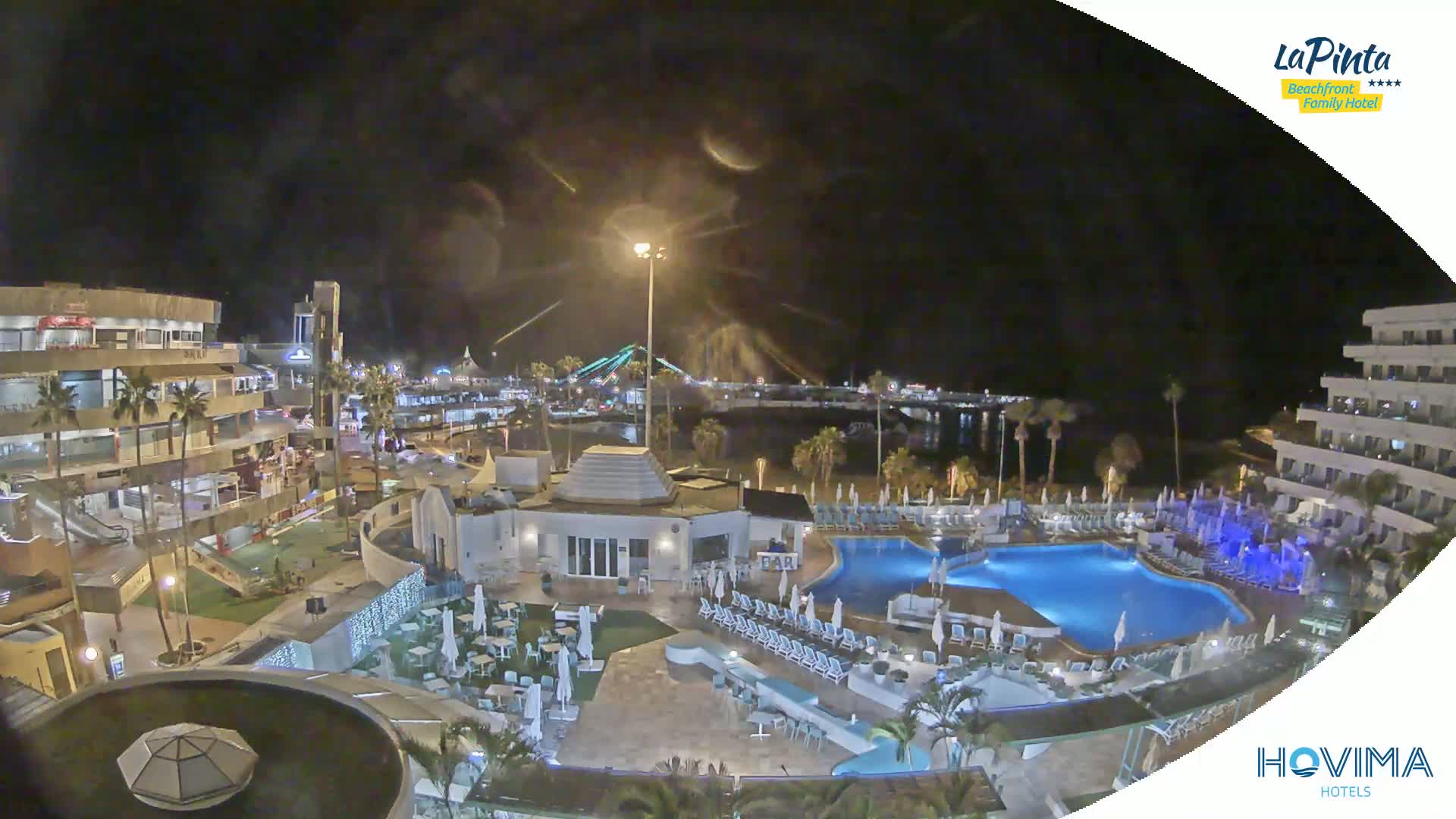 A nighttime view of a resort area with a swimming pool, surrounding buildings, and a waterfront in the distance under a partly cloudy sky.