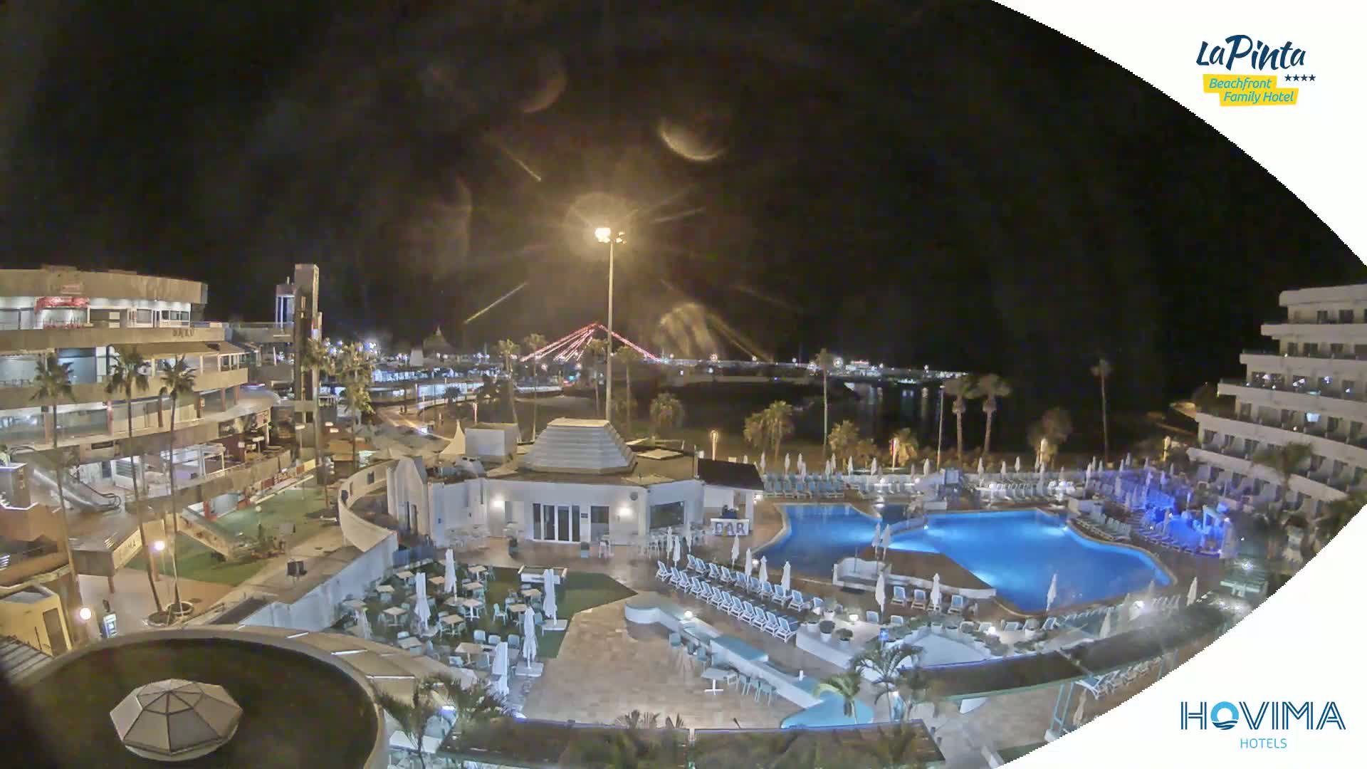 A nighttime aerial view of a resort complex with a swimming pool, surrounding buildings, and a bridge visible in the distance under a clear sky.