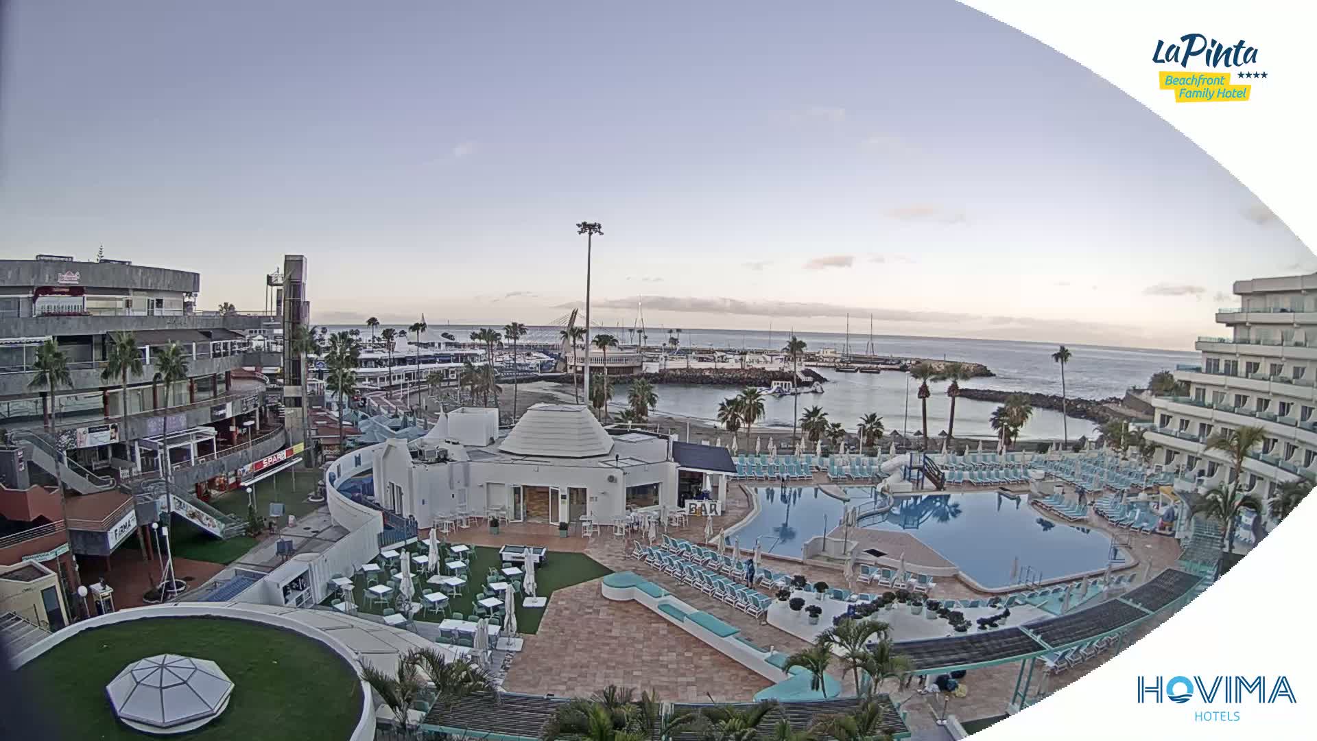 An aerial view shows a resort complex with a swimming pool, beach, and marina under a clear, twilight sky.