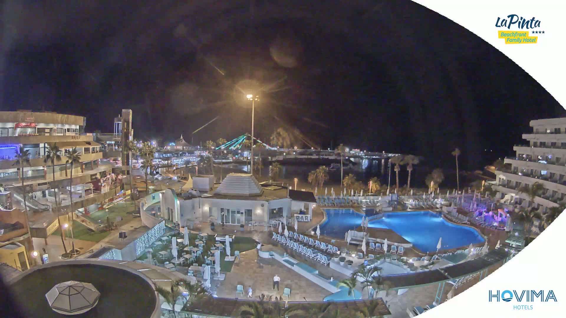 A nighttime aerial view of a resort complex with swimming pools, illuminated buildings, and a waterfront area under a dark sky.