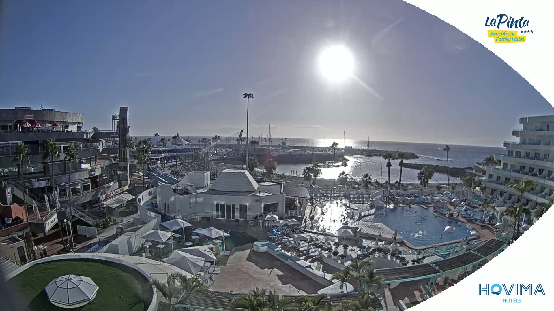 A sunny day overlooking a resort with a swimming pool, adjacent to a marina and shops near a calm ocean.