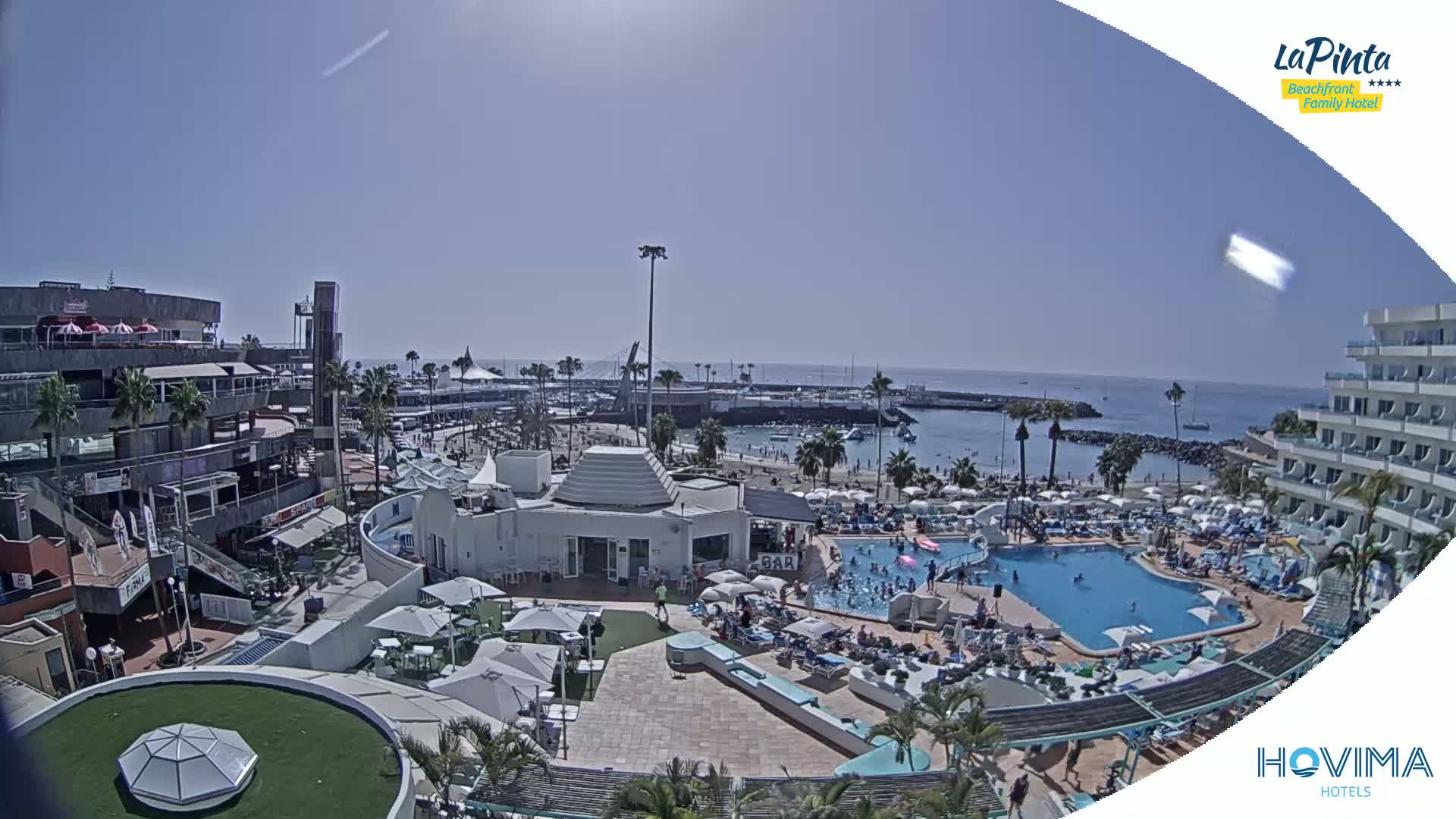 A sunny day overlooking a resort with a pool, beach, and harbor, showing buildings, palm trees, and people enjoying the sunny weather.