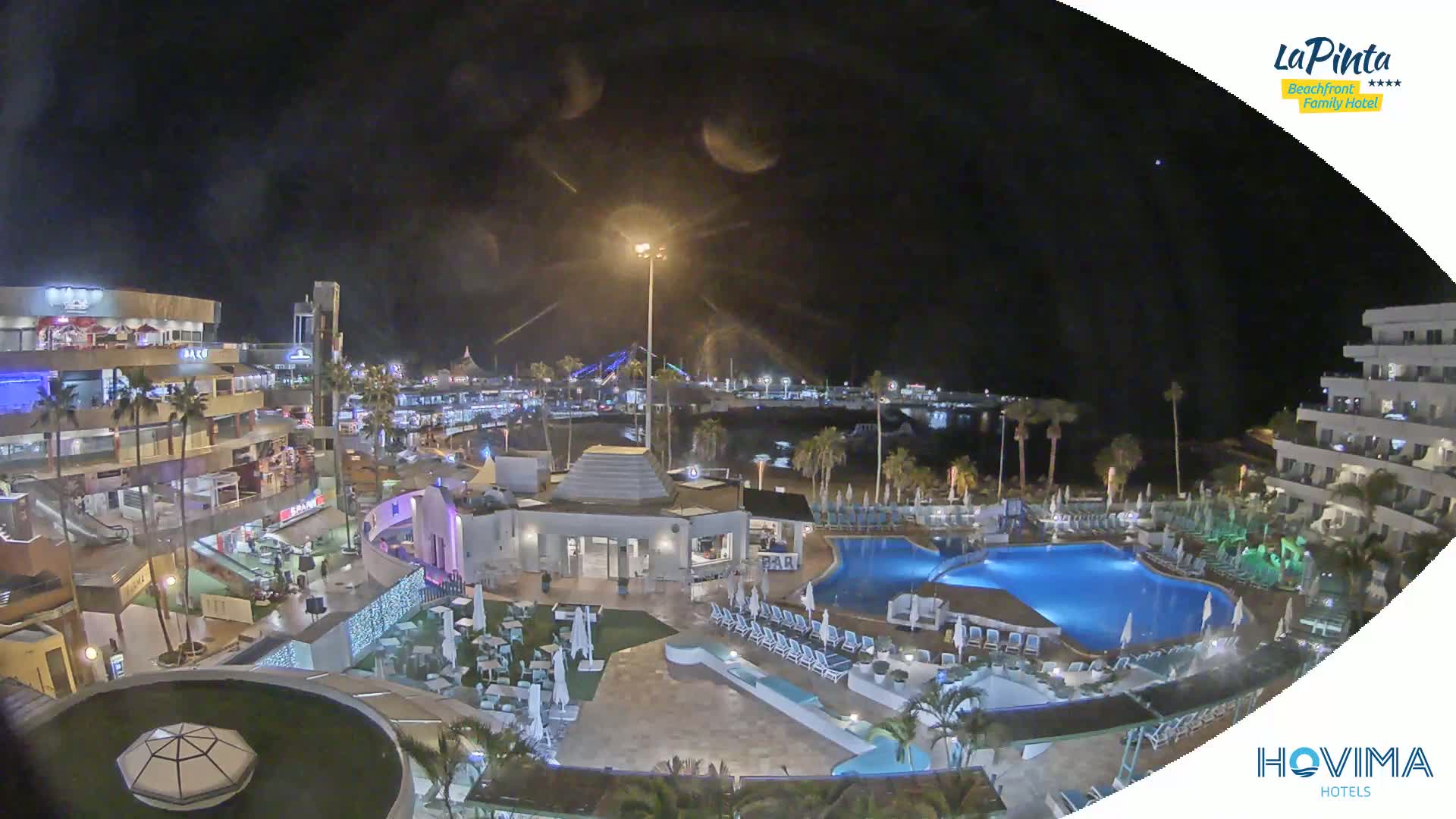 A nighttime aerial view of a resort complex with swimming pools, a plaza, and a marina in the background under a clear, dark sky.