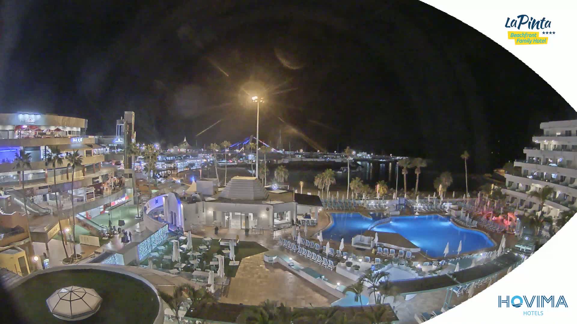 A nighttime aerial view of a resort complex with pools, buildings, and a beach, under a clear, dark sky.