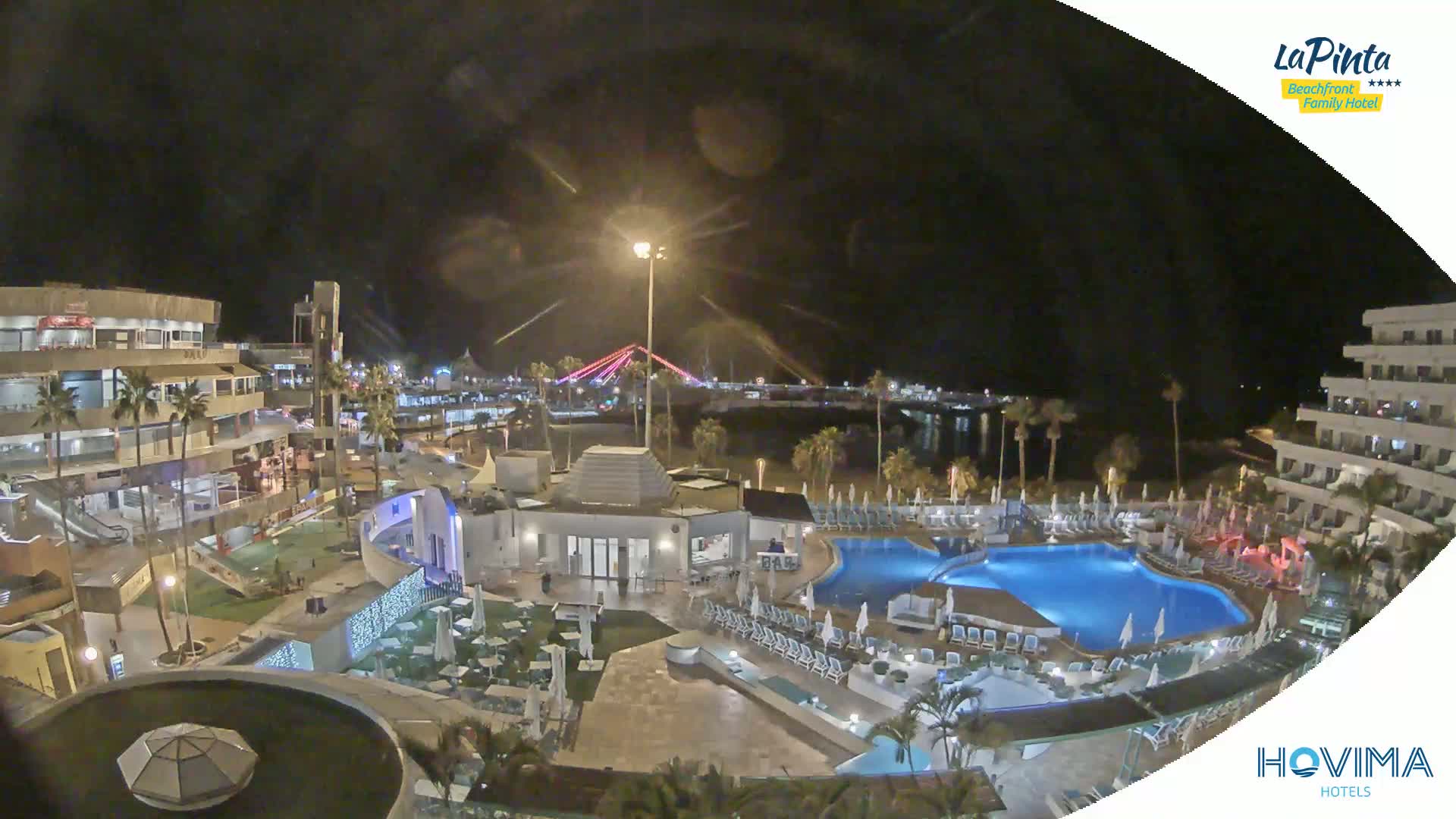 A nighttime aerial view of a resort with pools and surrounding buildings, illuminated under a dark sky.