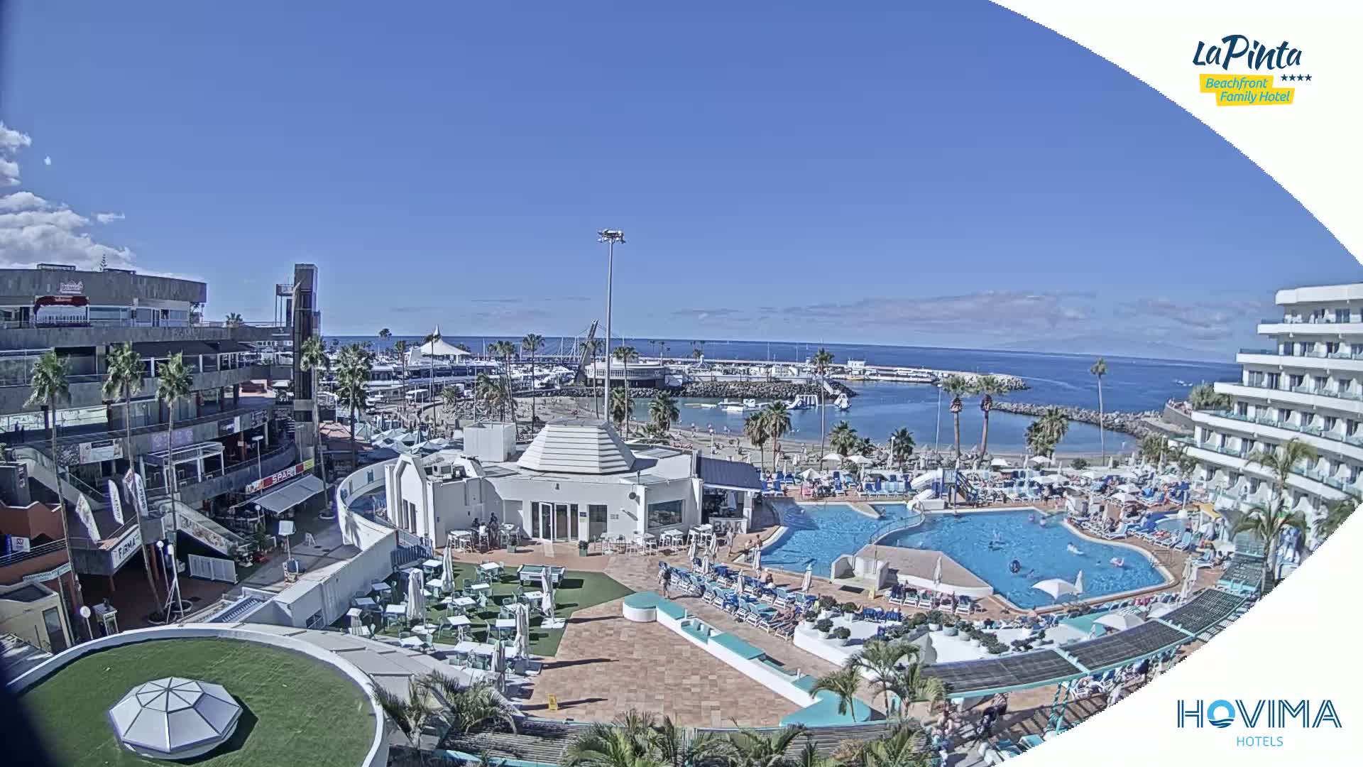 A sunny day overlooks a resort with a pool, beach, and marina,  adjacent to buildings and shops.