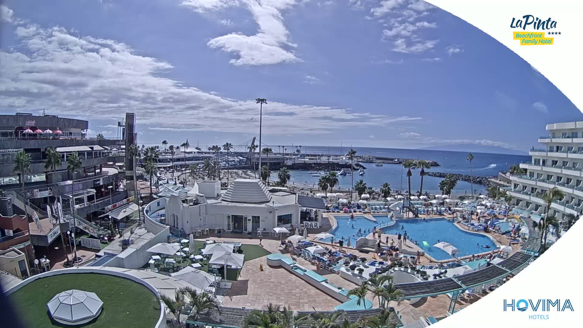 A sunny day overlooking a resort pool area, beach, and harbor, with partly cloudy skies.