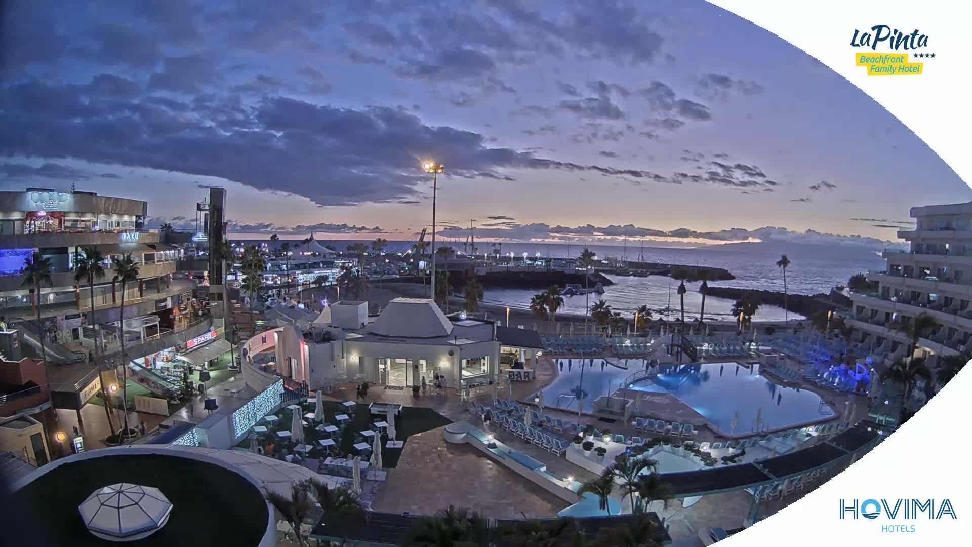 A resort pool area at dusk overlooks a calm ocean harbor and a waterfront shopping district under a partly cloudy sky.