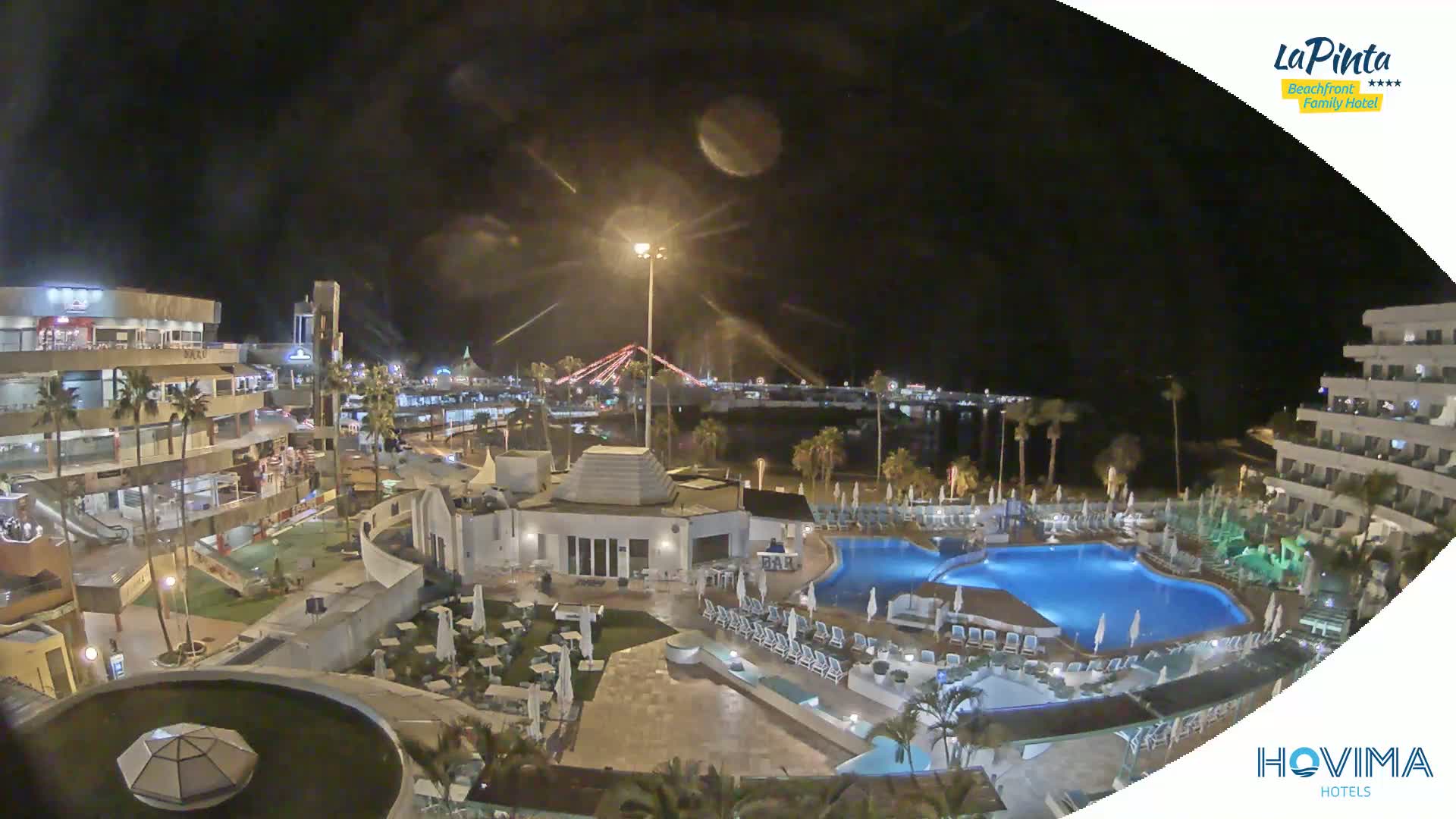 A nighttime aerial view of a resort complex with a lit-up swimming pool, surrounding buildings, and palm trees, overlooking a calm body of water and a bridge in the distance under a clear, dark sky.