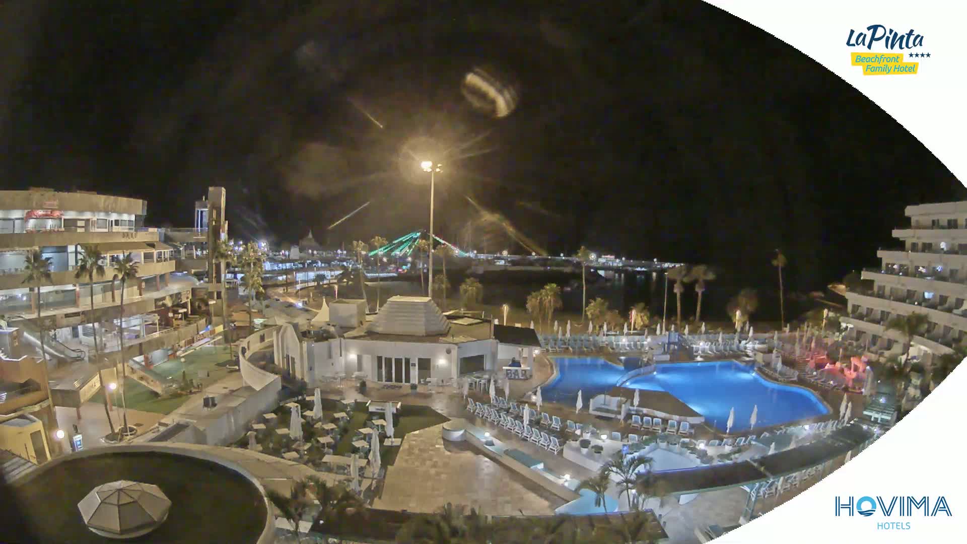 A nighttime aerial view of a resort complex with pools and surrounding buildings, under a dark, clear sky.