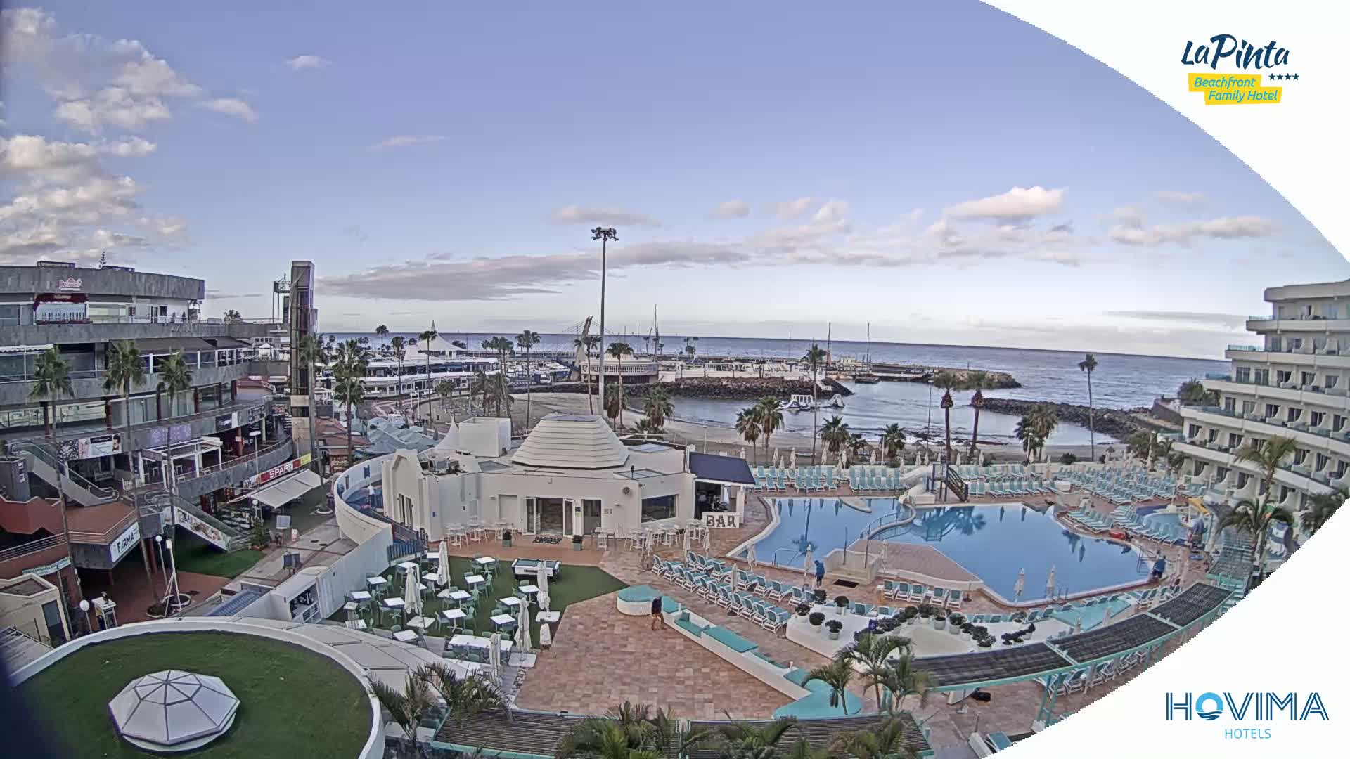An outdoor scene shows a resort area with a swimming pool, shops, and a marina under a partly cloudy sky.