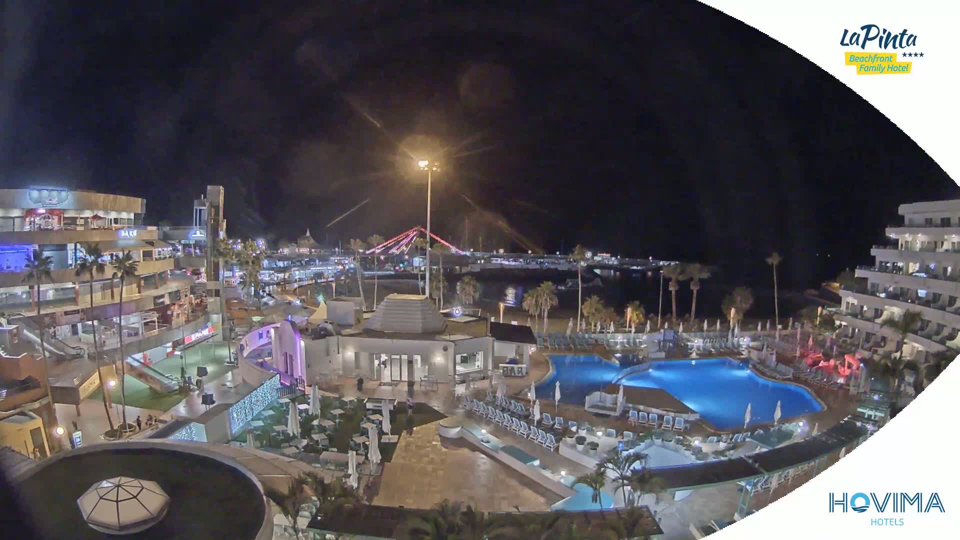 A nighttime aerial view of a resort complex with illuminated buildings, palm trees, and a swimming pool, overlooking a beach and a bridge.