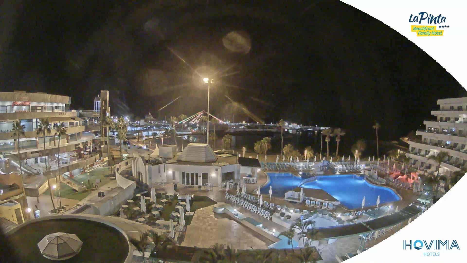 A nighttime aerial view of a resort complex with a swimming pool, surrounding buildings, and a waterfront illuminated with lights.