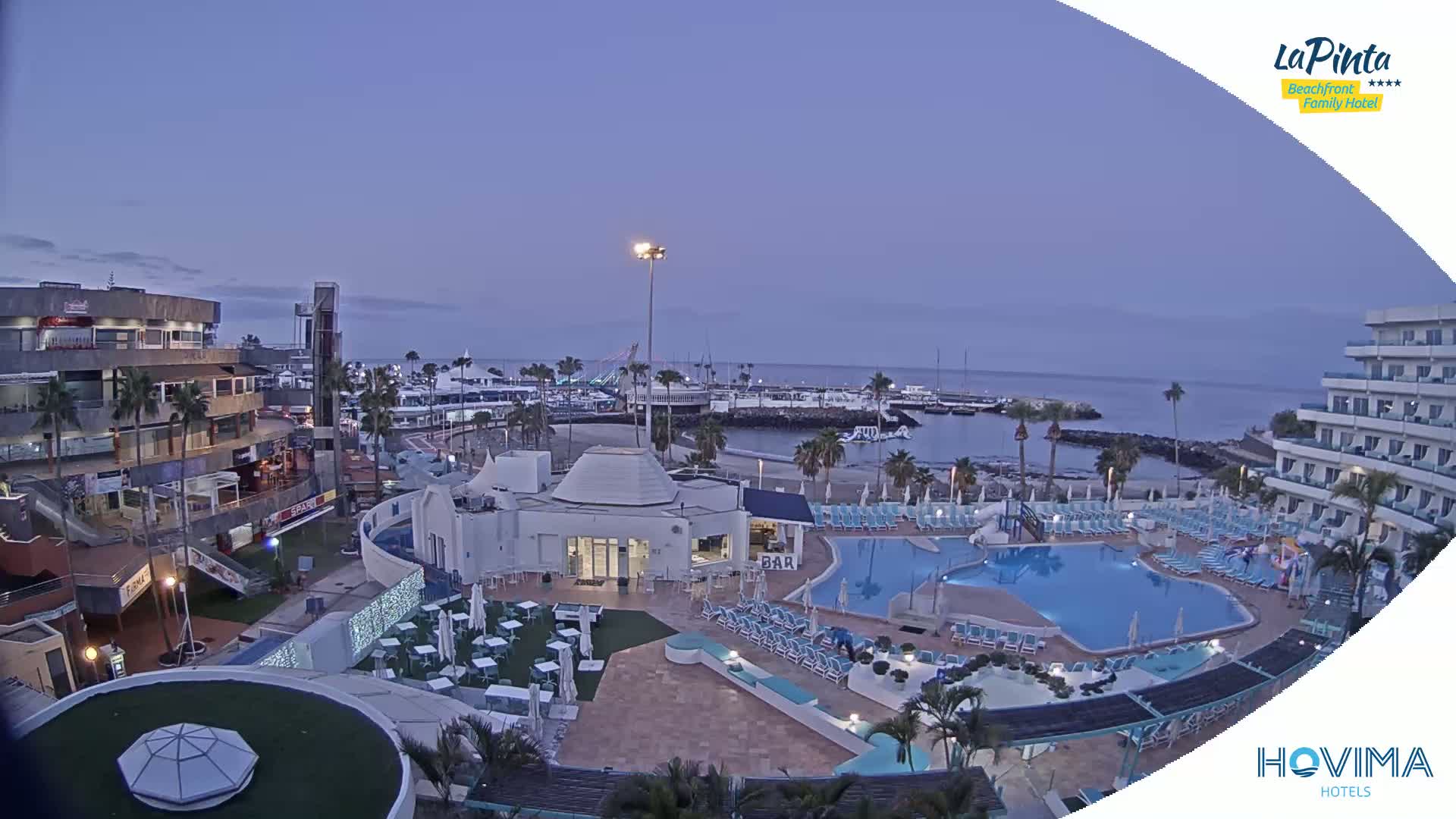 An elevated view of a resort area at dusk, showing a pool, buildings, a marina, and calm ocean water.