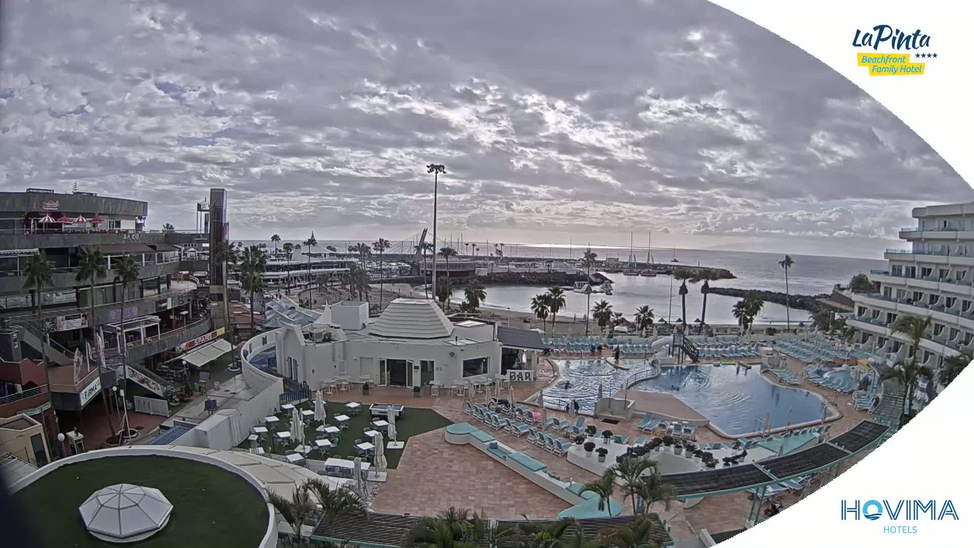 An overcast day reveals a resort scene with a swimming pool, beach, marina, and shops, all situated near a calm ocean.