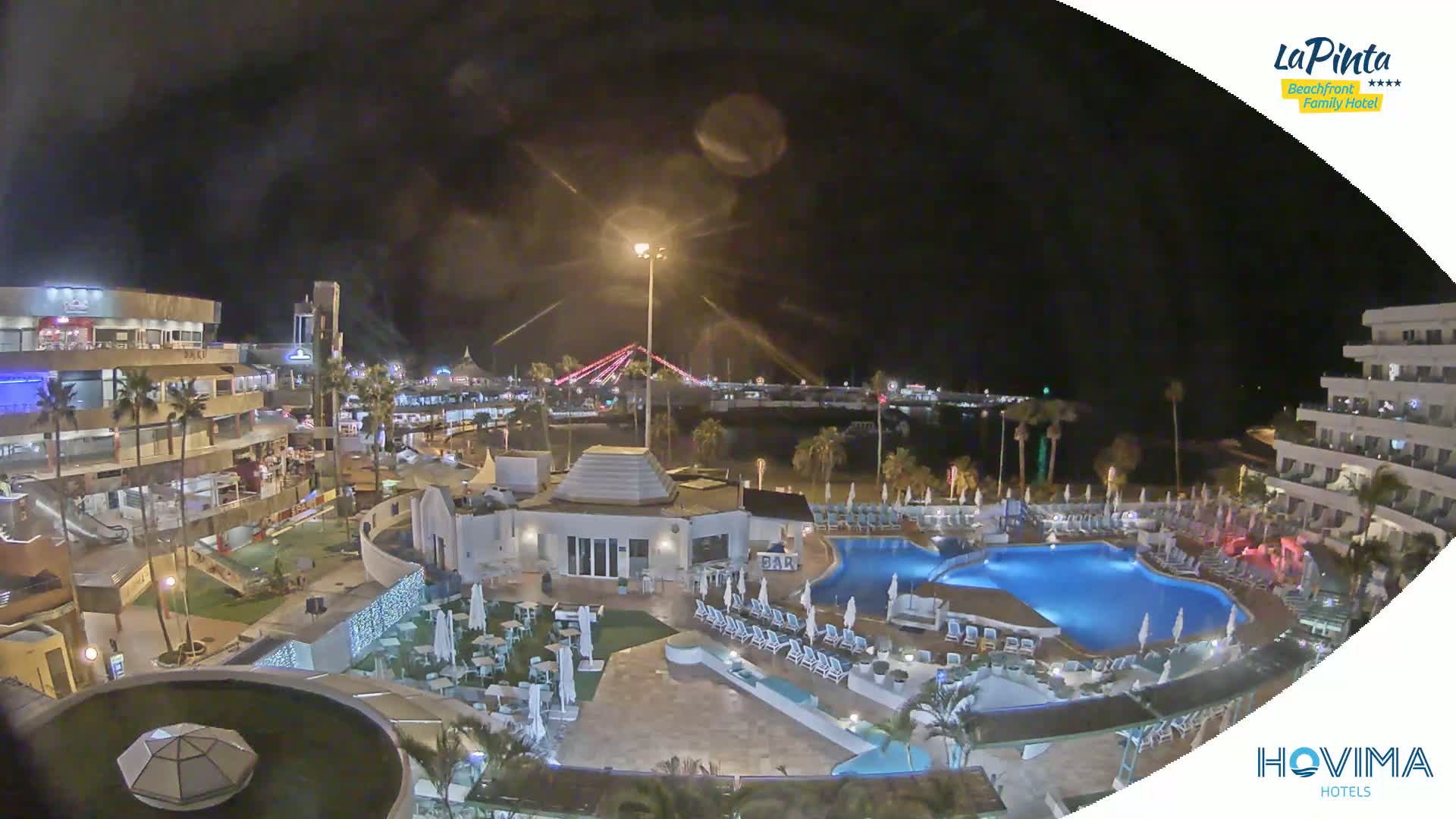 A nighttime aerial view of a resort complex with multiple pools, surrounding buildings, and a beach in the background under a dark, clear sky.