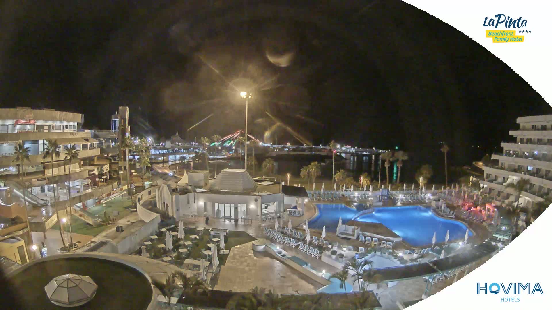 A nighttime aerial view shows a resort complex with a pool area, surrounding buildings, and a calm body of water in the background under a dark, clear sky.
