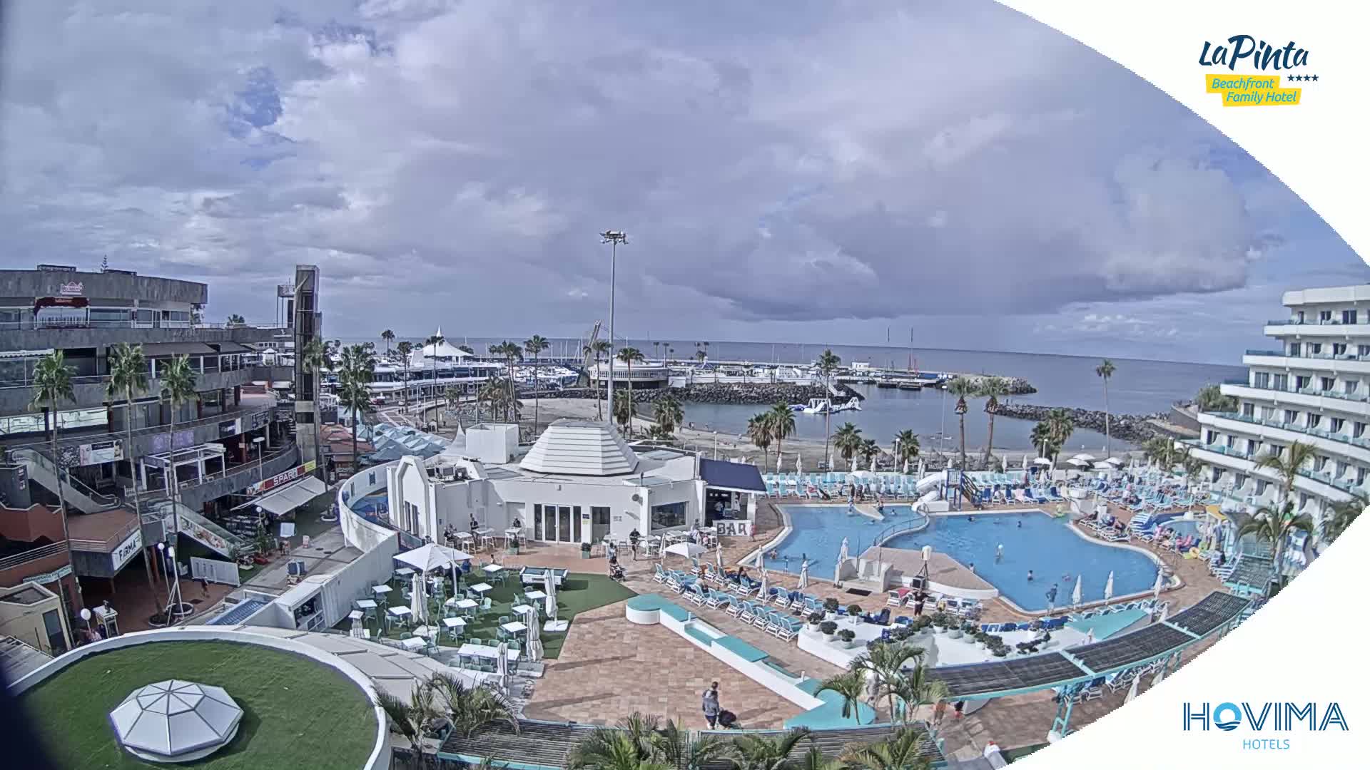 An outdoor view of a resort complex with a swimming pool, a marina, and buildings under a partly cloudy sky.