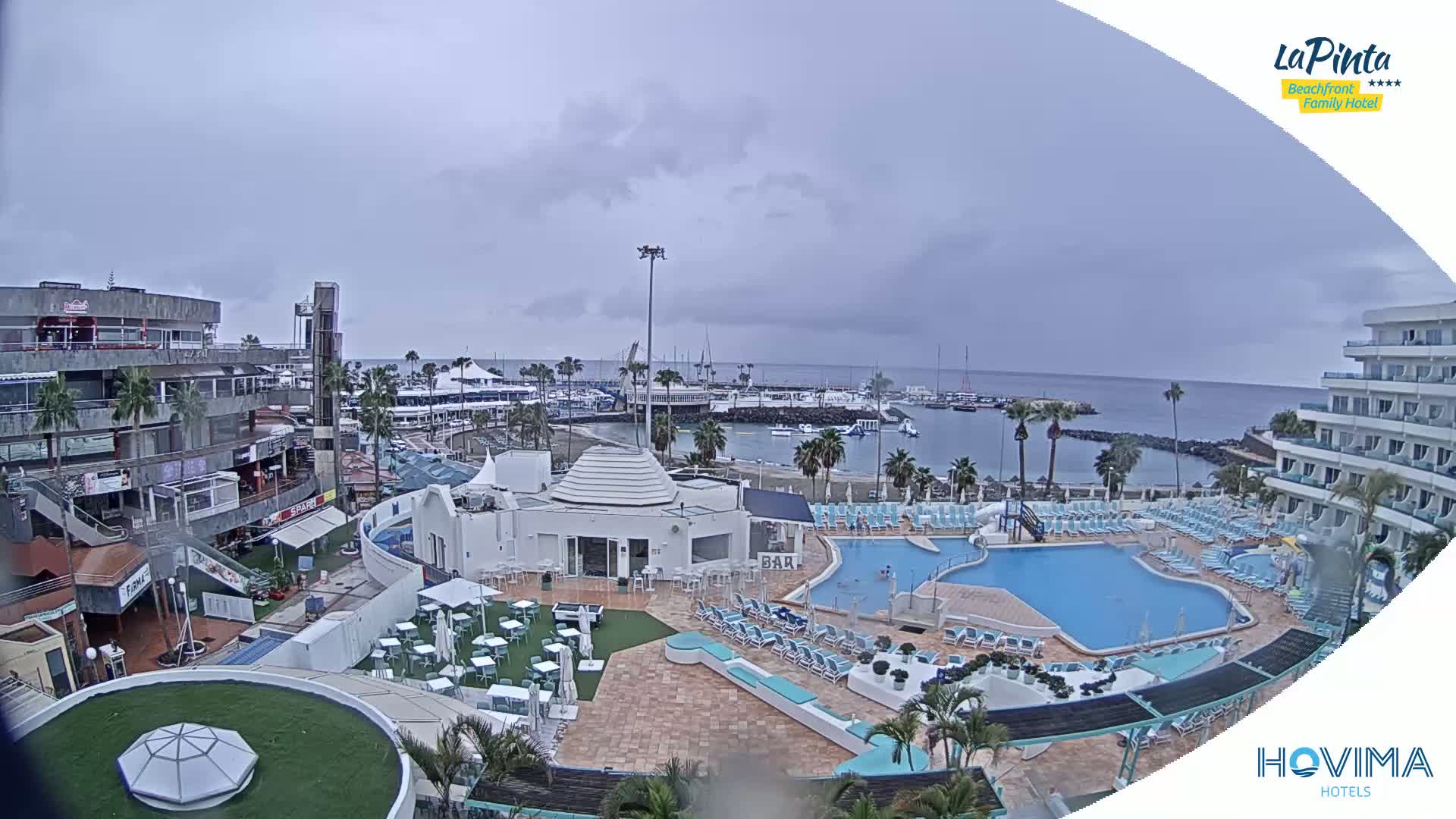 An overcast day shows a resort with a pool and a marina, situated near a calm ocean.