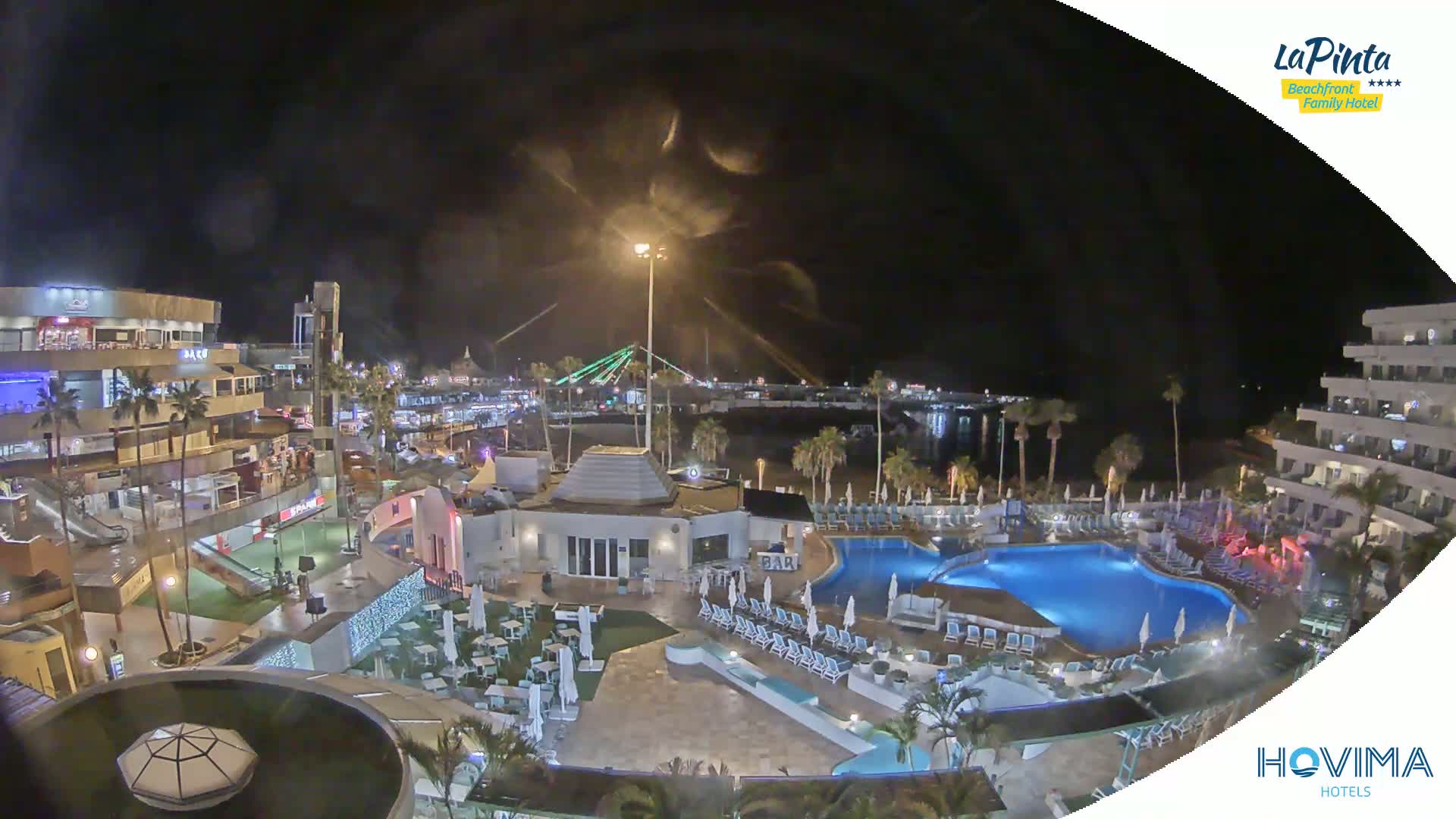 A nighttime aerial view of a resort complex with illuminated pools, buildings, and walkways, overlooking a calm body of water and a bridge.