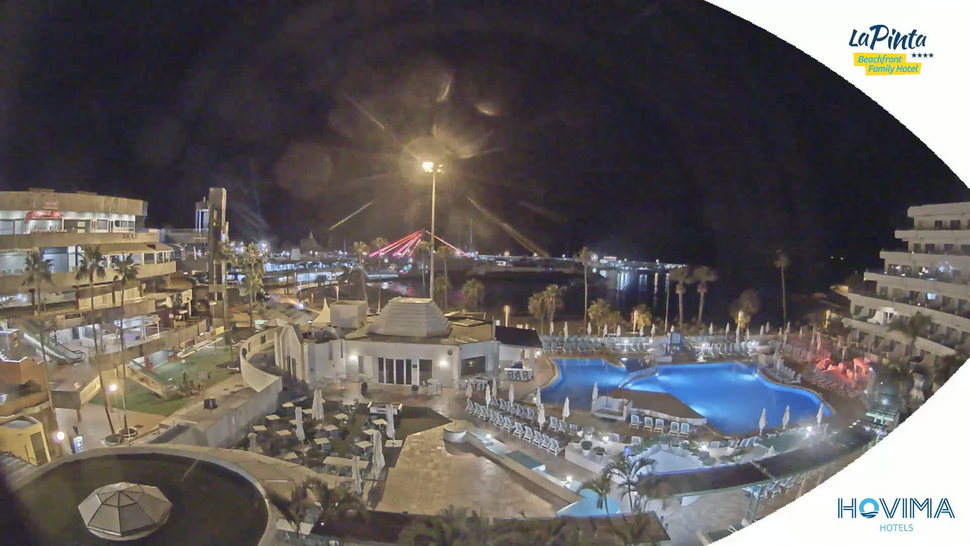 A nighttime aerial view of a resort complex with swimming pools and palm trees, overlooking a harbor and a lit bridge under a dark, clear sky.