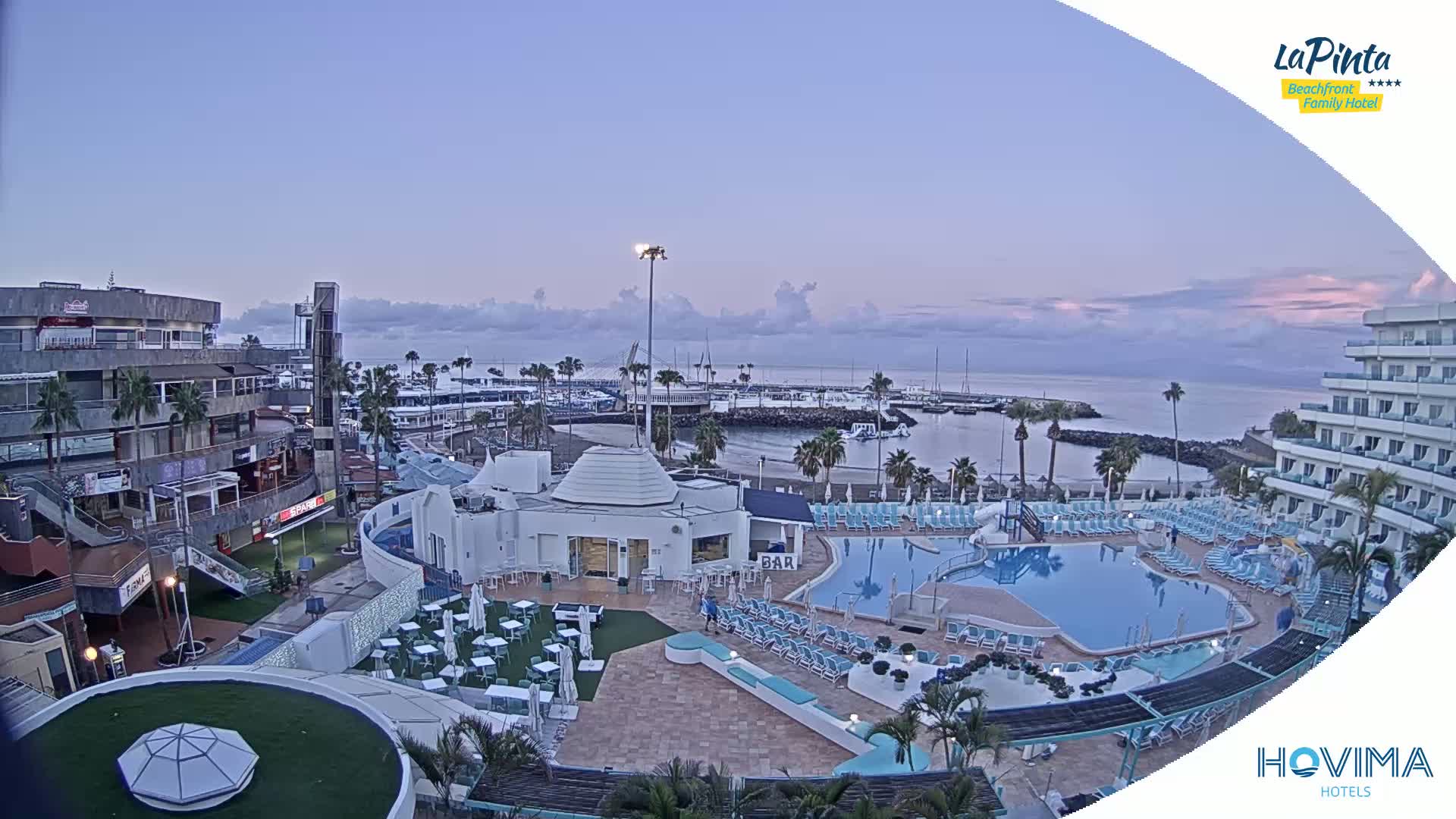An aerial view of a resort with a swimming pool overlooking a calm harbor at dawn under a mostly clear sky.
