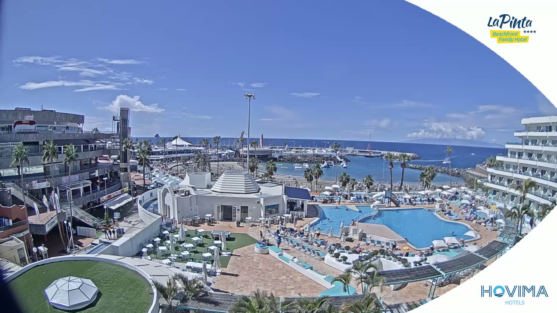 A sunny day overlooks a resort with a pool and beach access near a marina and shops.