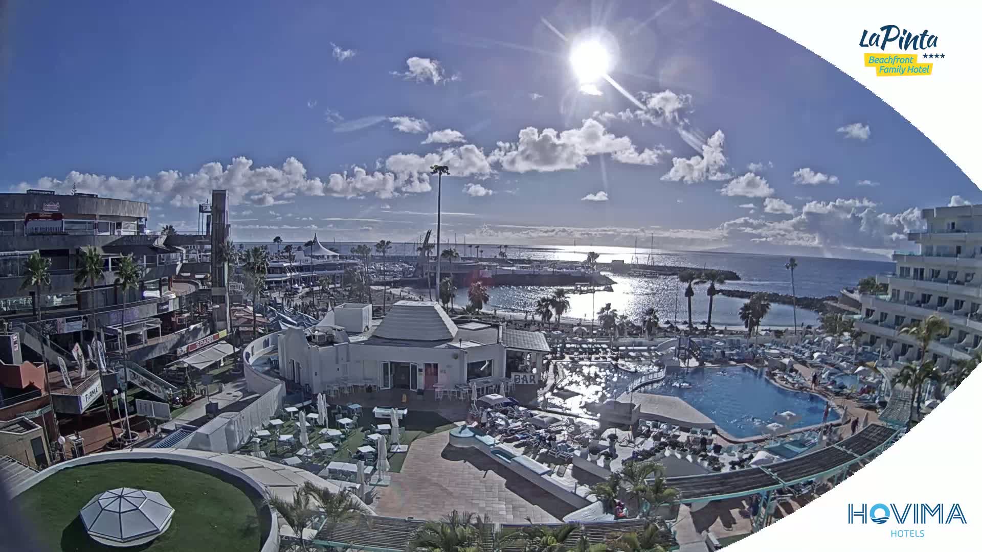 A sunny day reveals a resort area with a swimming pool, restaurants, shops, and a marina, all overlooking a calm ocean.