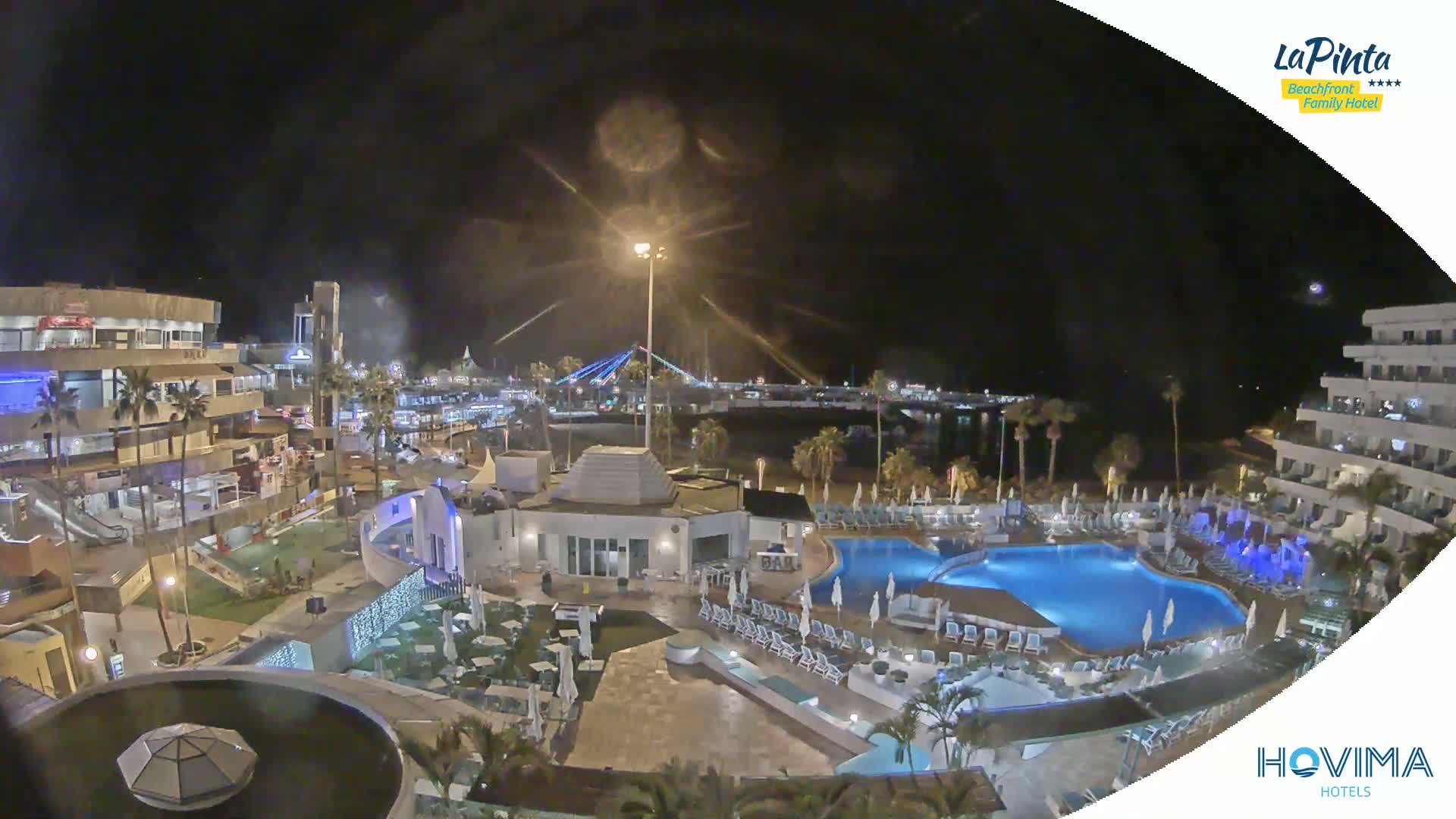 A nighttime aerial view of a resort complex with pools and surrounding buildings, illuminated under a dark sky.