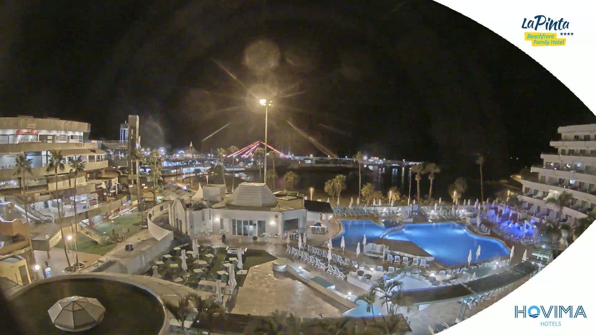 A nighttime aerial view of a resort with swimming pools, illuminated buildings, and palm trees, overlooking a calm body of water and a bridge in the distance.