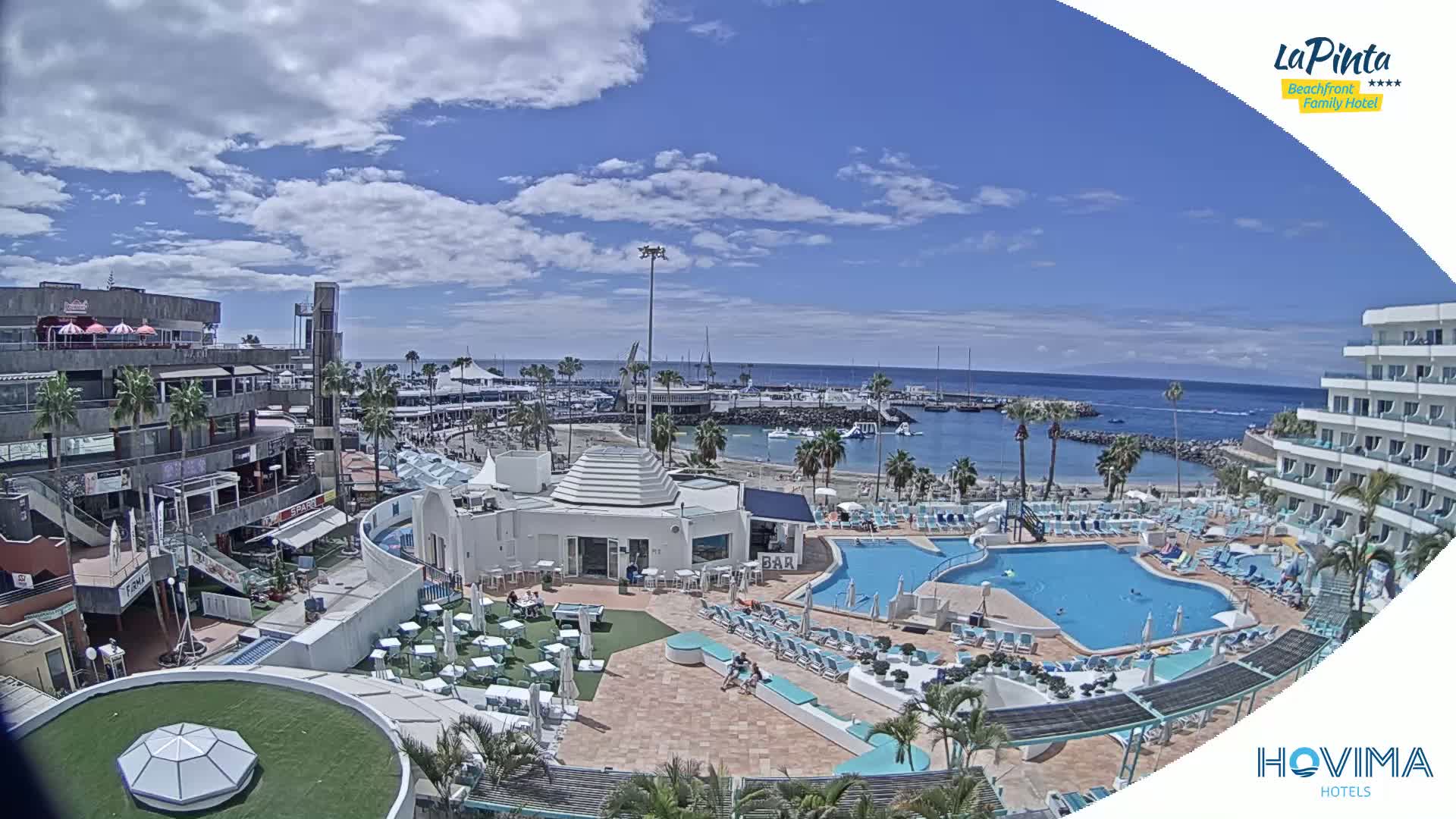 A sunny day overlooking a resort with multiple pools, a marina, and a shopping area.