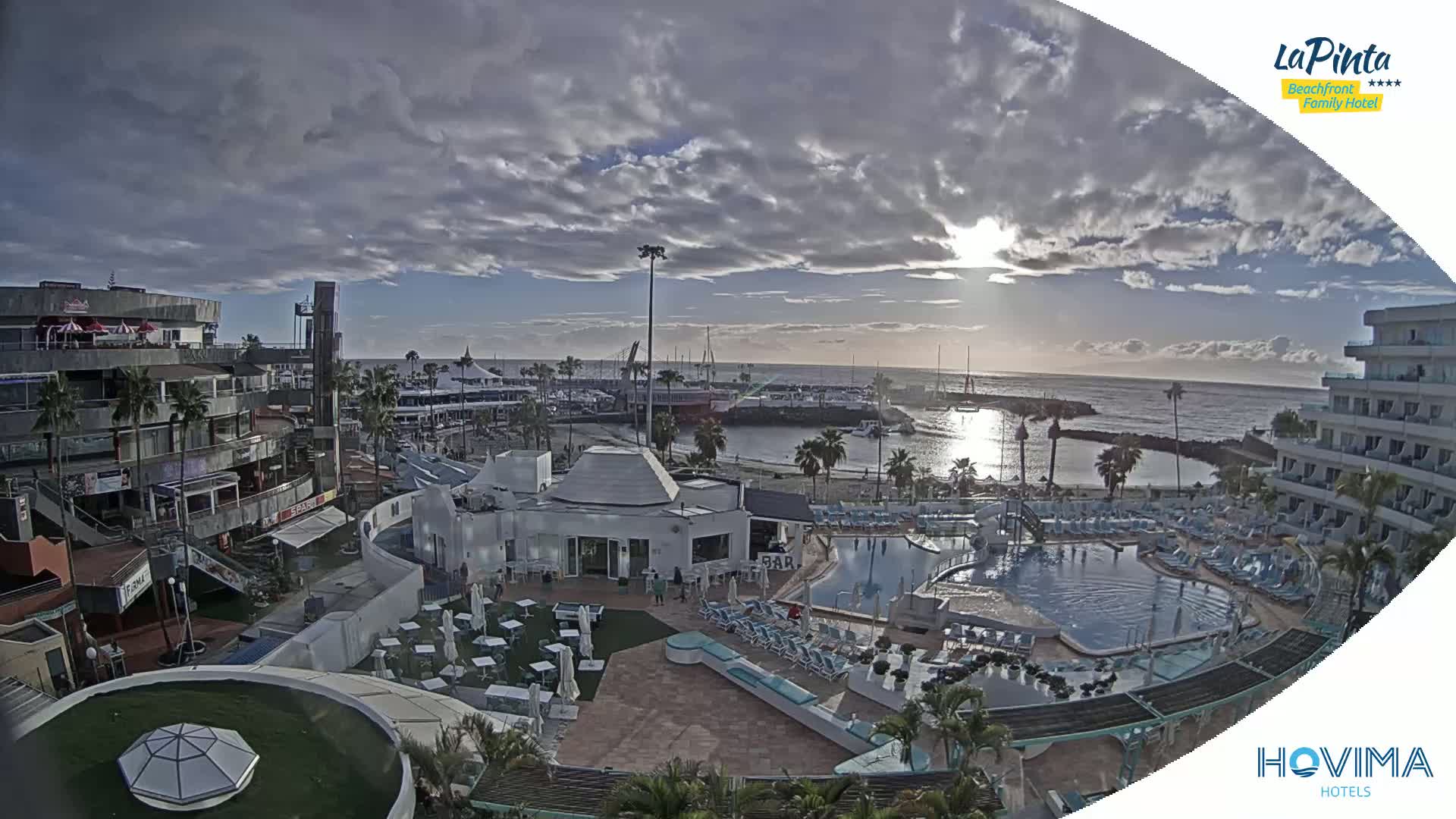 An aerial view of a resort complex with a swimming pool, situated near a harbor and ocean under a partly cloudy sky.