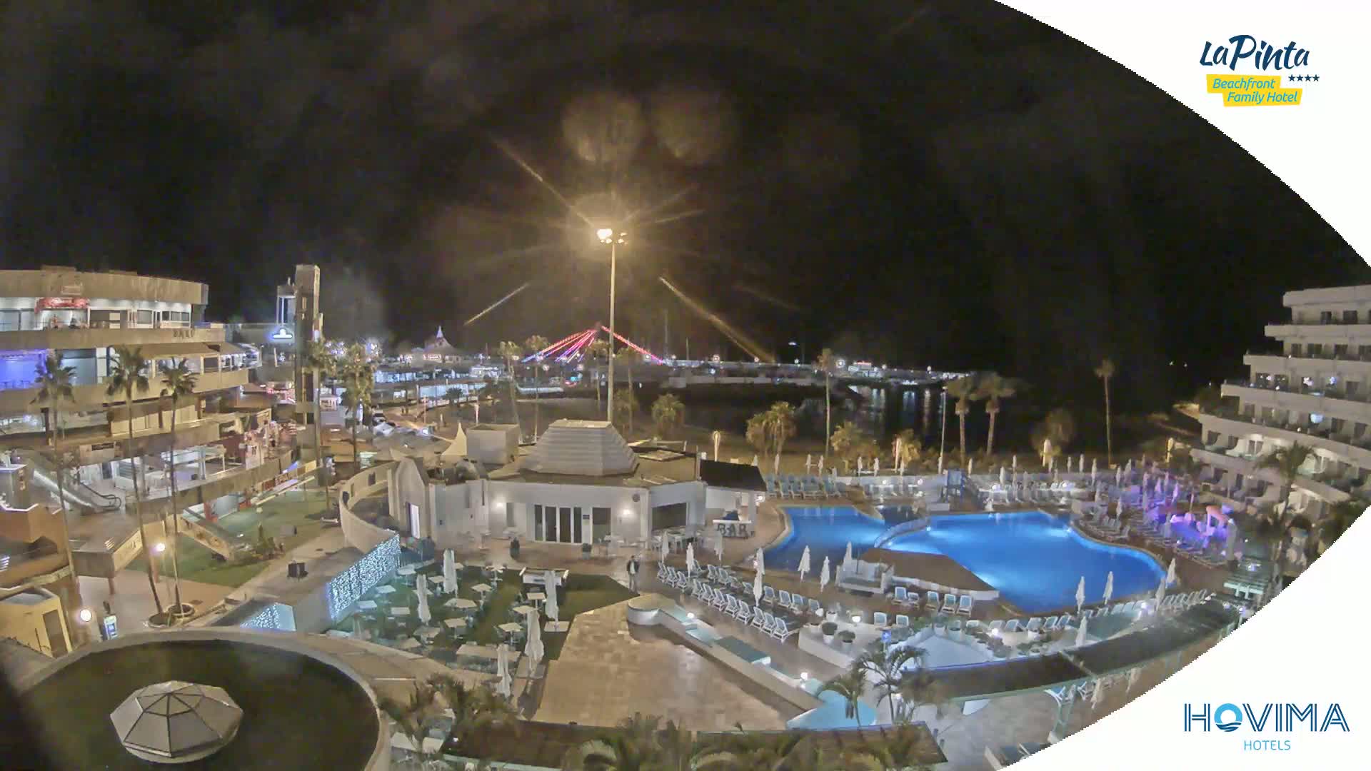 A nighttime aerial view shows a resort complex with a swimming pool, surrounding buildings, and a beach with a bridge in the distance under a dark sky.