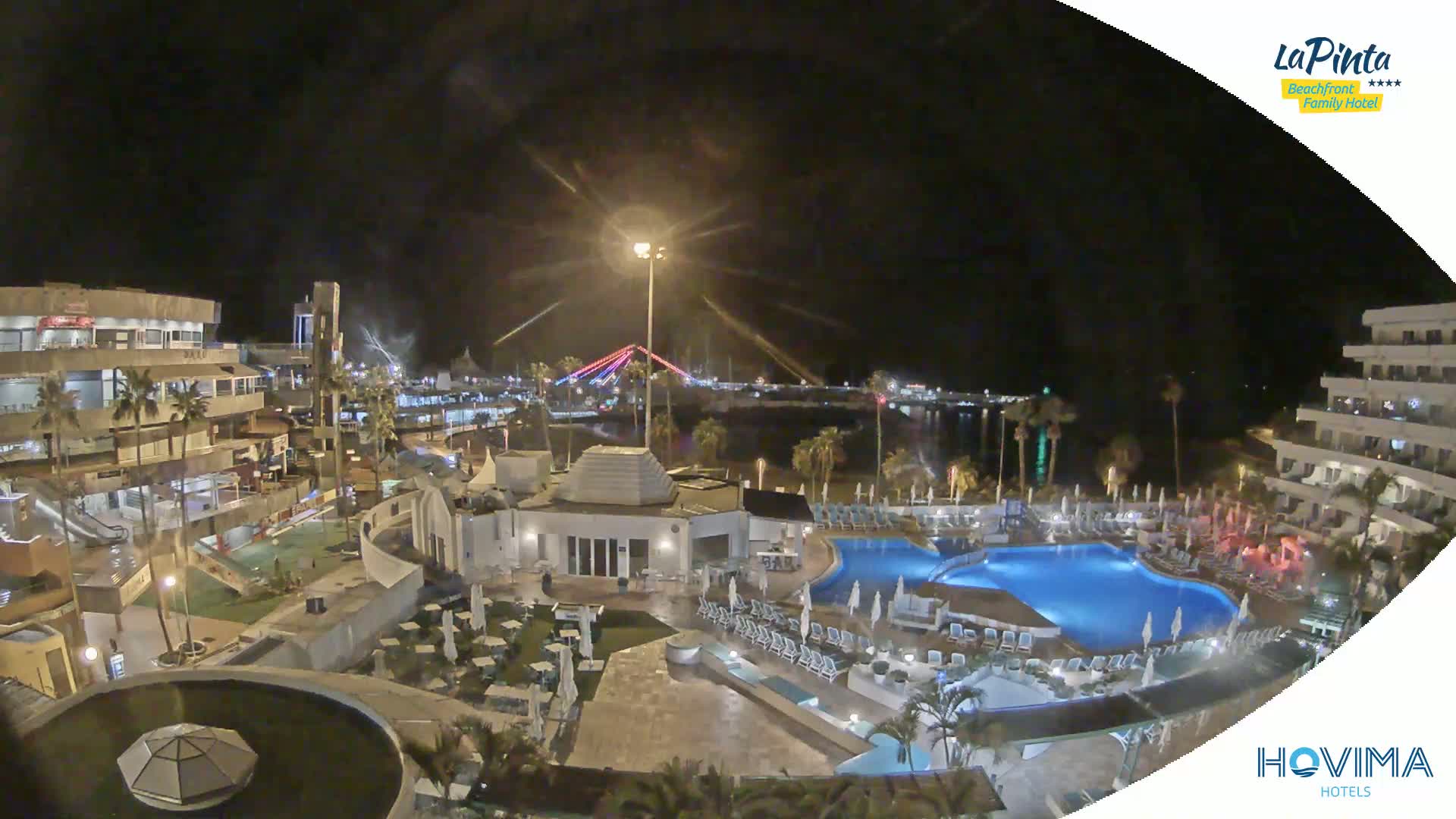 A nighttime view of a resort complex with a swimming pool, situated near a body of water and a lit-up bridge under a dark sky.