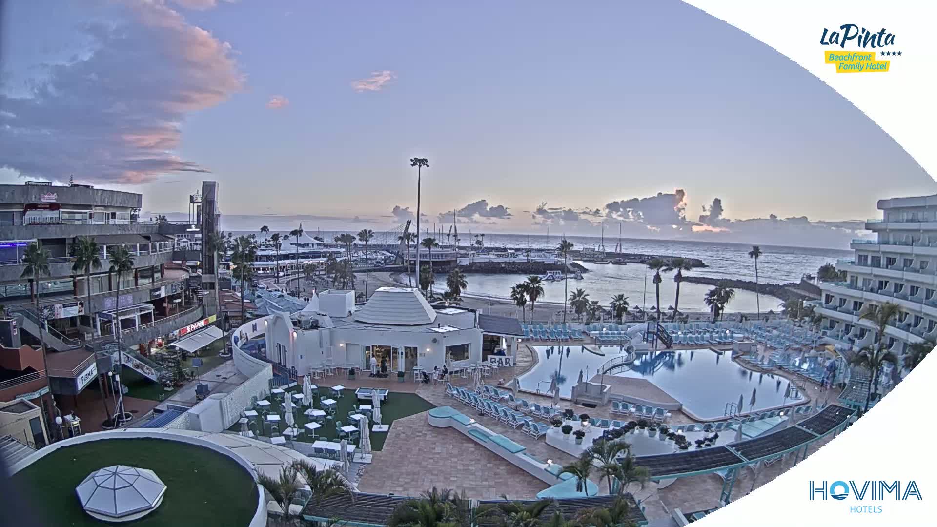 An elevated view shows a resort area with a swimming pool, beach, marina, and buildings under a mostly clear twilight sky.