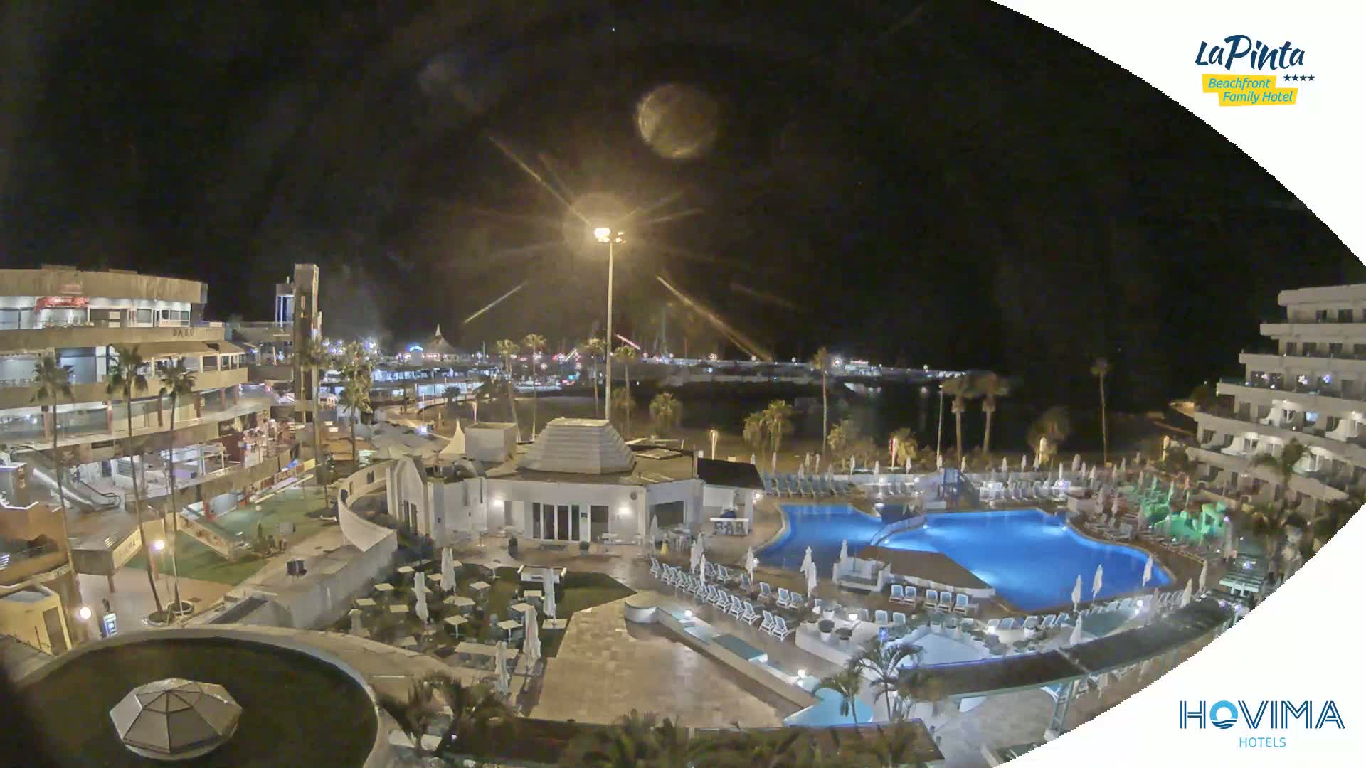 A nighttime aerial view of a resort complex with pools and surrounding buildings, under a partly cloudy sky and bright moonlight.