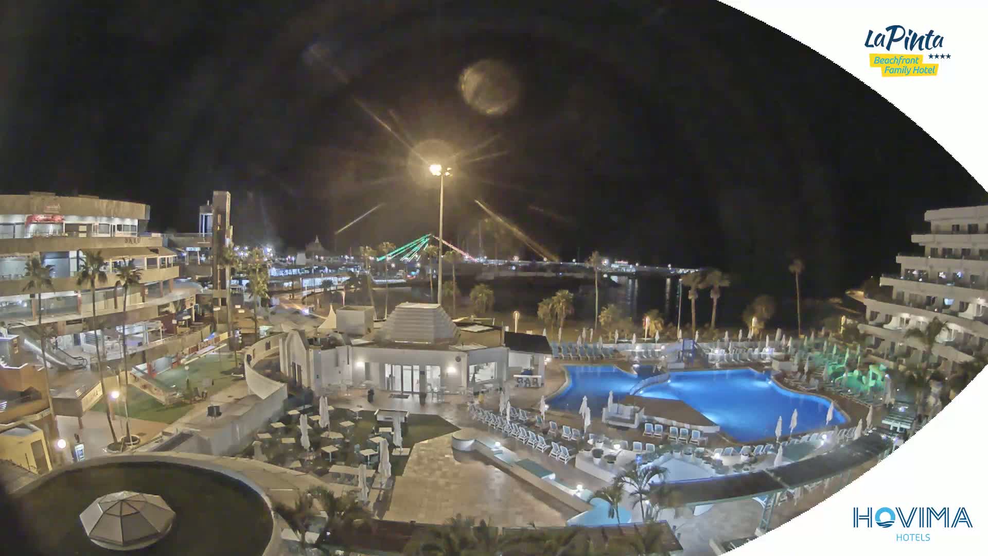 A nighttime aerial view of a resort complex with multiple pools, surrounding buildings, and a waterfront with a bridge in the distance under a clear, dark sky.