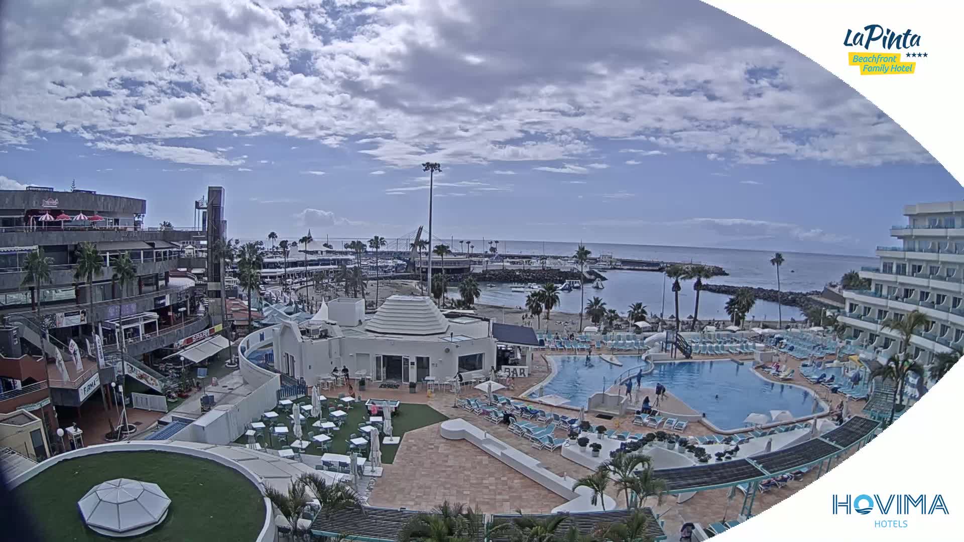 A resort pool area with a small beach and marina visible in the background under a partly cloudy sky.