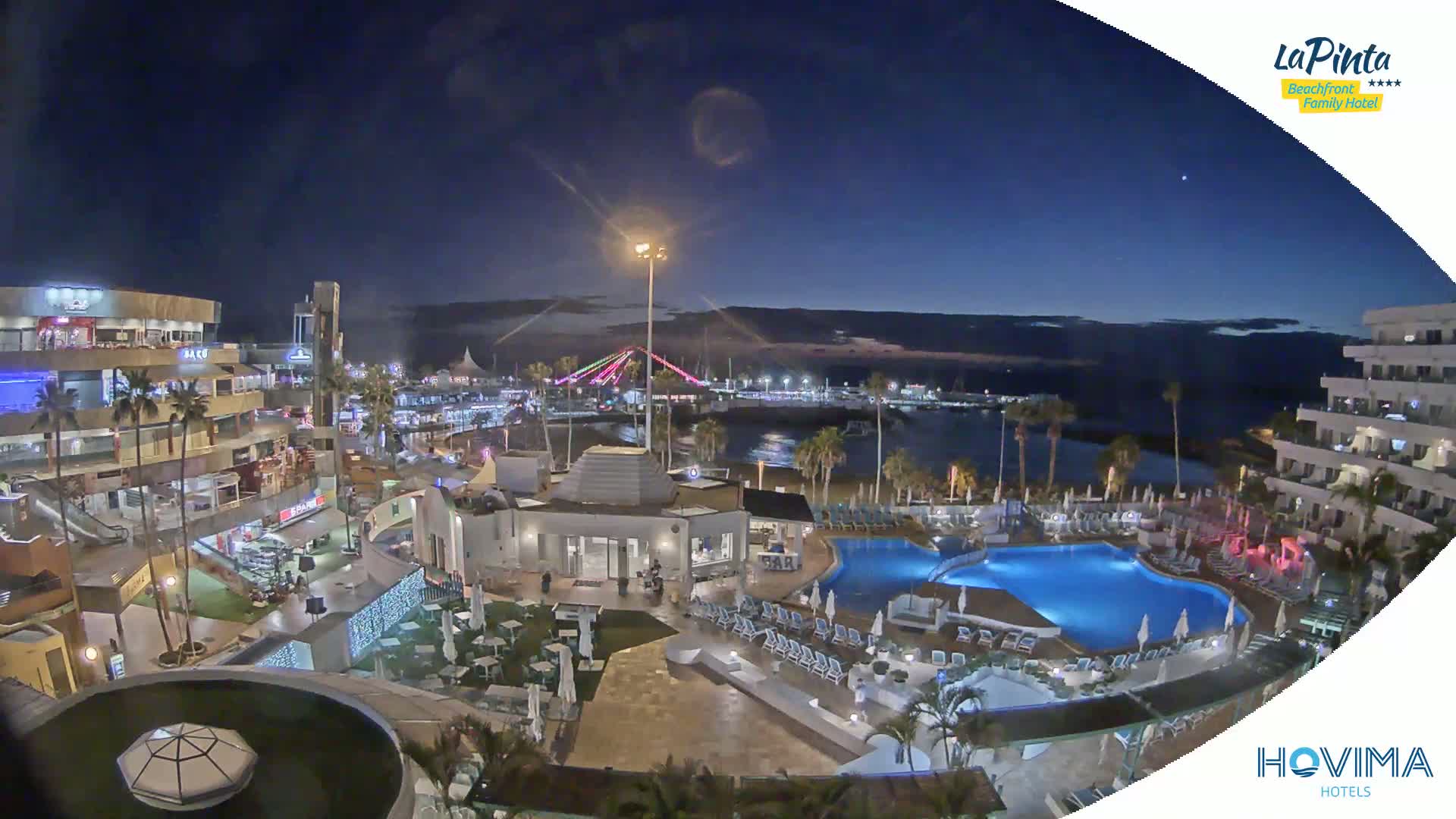 A nighttime aerial view shows a resort area with a swimming pool, buildings, and palm trees next to a calm body of water under a clear, dark sky.