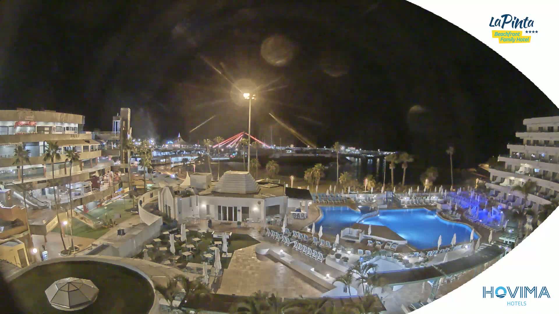 A nighttime aerial view of a resort with a swimming pool complex, illuminated buildings, and a beach, overlooking a body of water and a bridge in the distance under a clear, dark sky.
