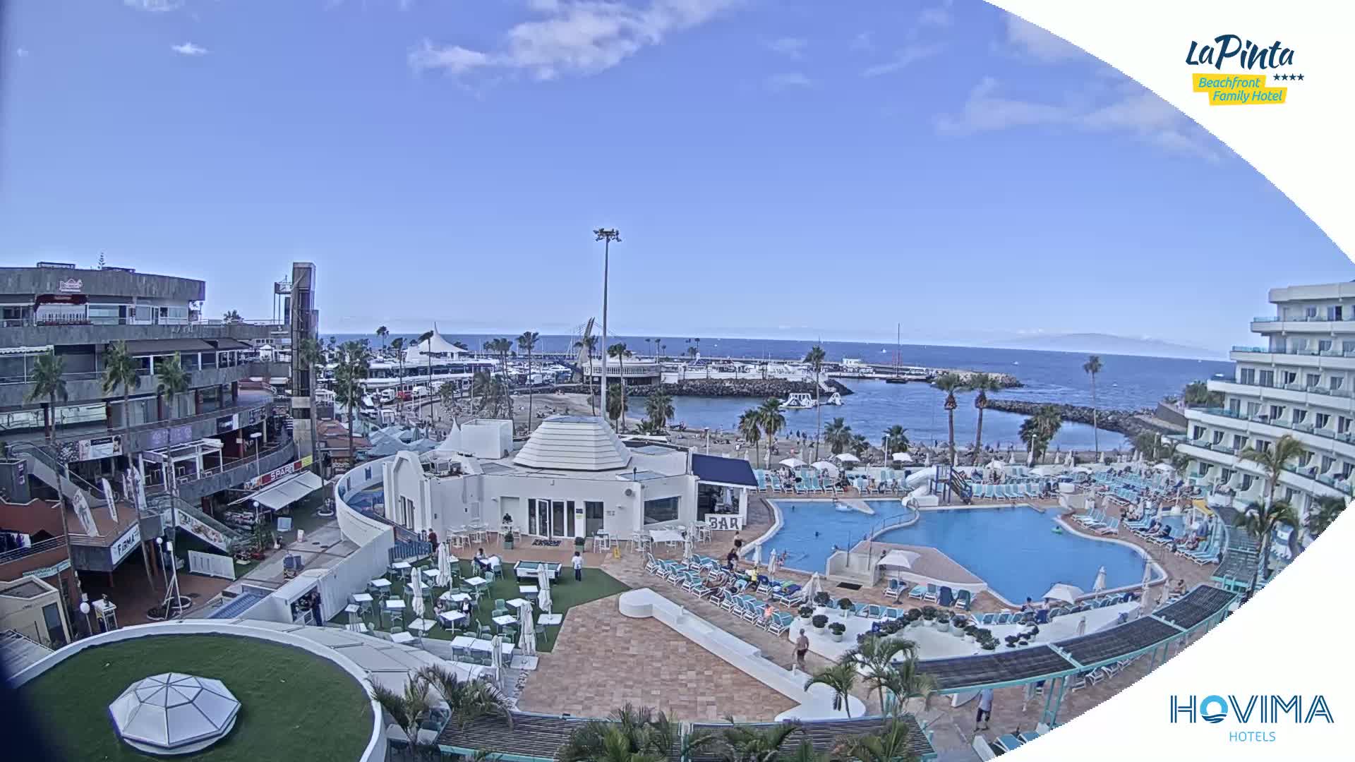 An aerial view of a resort area on a sunny day shows a swimming pool, beach, marina, and buildings.