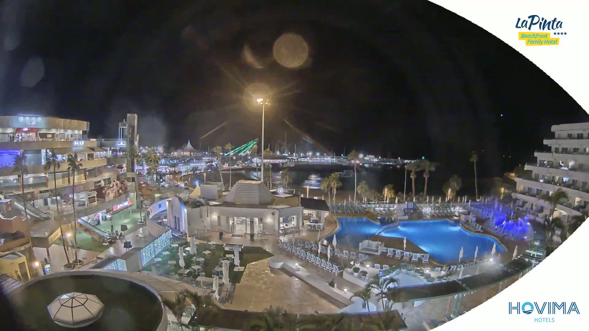 A nighttime aerial view of a resort complex with pools and surrounding buildings, overlooking a calm harbor under a dark, clear sky.