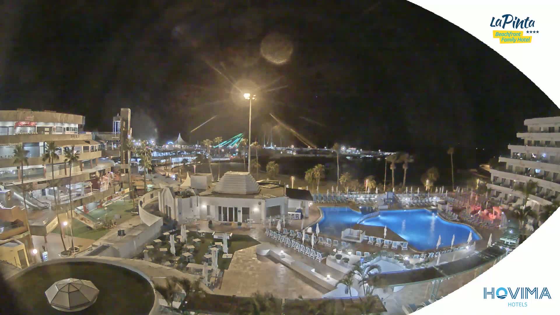 A nighttime aerial view of a resort complex with a pool, surrounding buildings, and a beachfront area beyond.