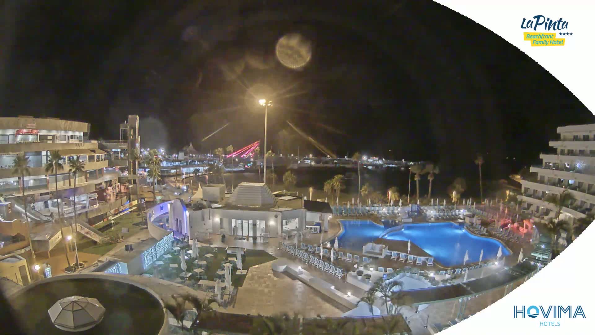 A nighttime aerial view of a resort complex with a swimming pool, situated near a waterfront area with lights illuminating the scene.