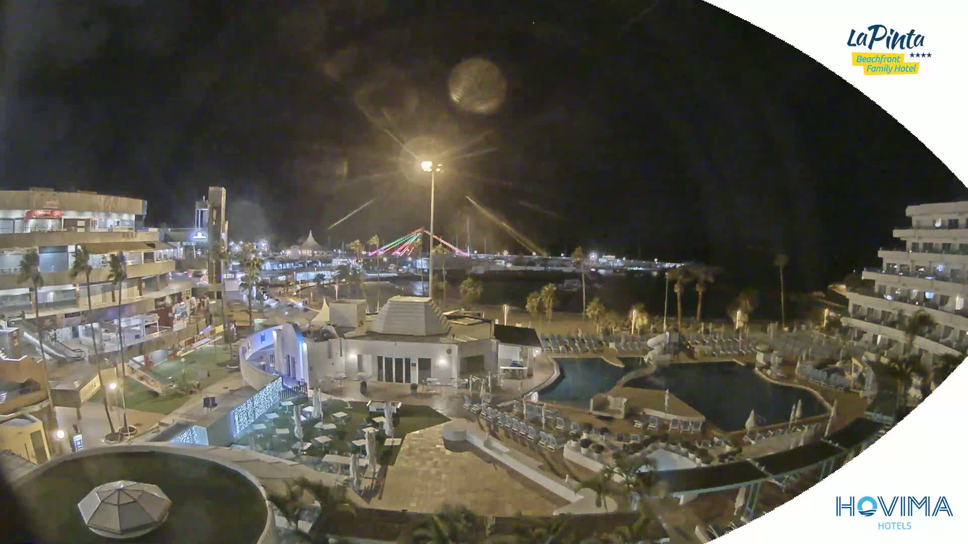 A nighttime aerial view of a resort complex with swimming pools, buildings, and landscaping, overlooking a beach and a brightly lit bridge.