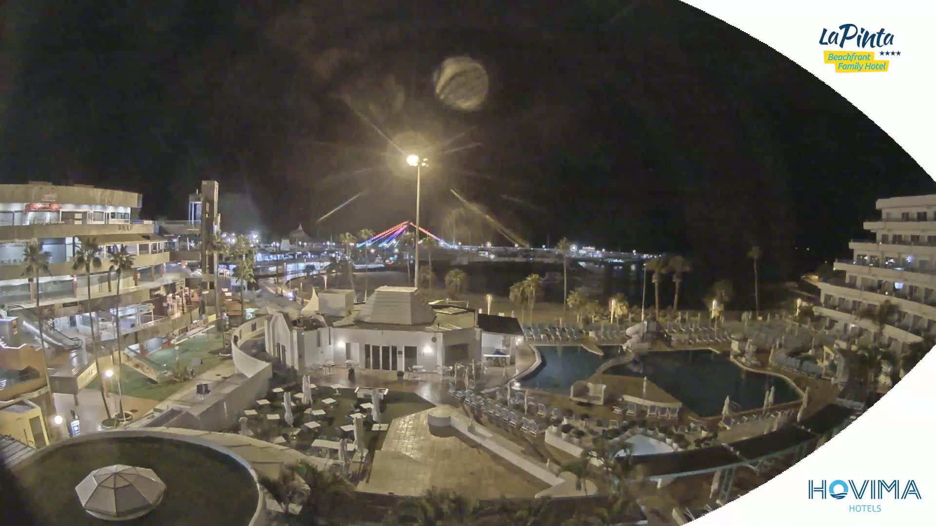 A nighttime aerial view of a resort complex with pools and outdoor seating areas, overlooking a beach and illuminated bridge under a clear, dark sky.
