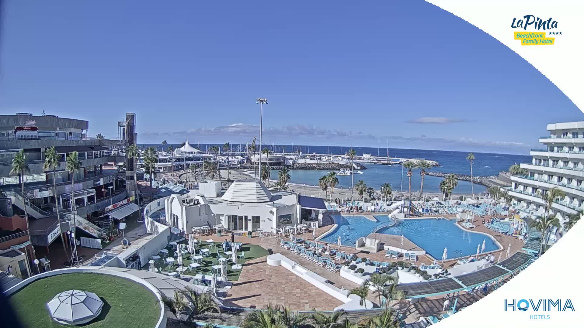 An aerial view shows a resort complex with a swimming pool, adjacent to a marina and beach under sunny skies.
